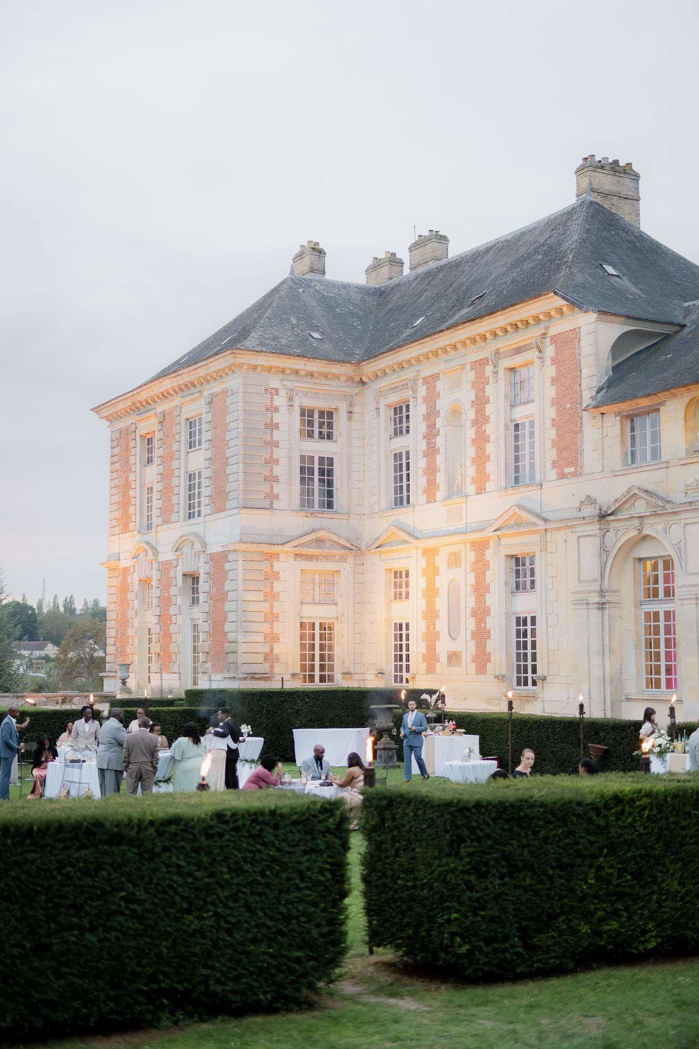 An outdoor cocktail hour or reception taking place in the formal garden grounds of a French baroque-style chateau, photographed at dusk from a wide-angle, elevated perspective. The chateau features red brick and cream stone facade with a slate mansard roof, and its exterior is warmly lit by uplighting. Approximately 20–25 guests are gathered around white-clothed cocktail tables and seated arrangements between manicured box hedgerows, with tall torch-style flame lights positioned throughout the garden space. Guests are dressed in formal attire including suits and occasion wear in neutral and muted tones. The composition is taken from behind a gap in the trimmed hedges, framing the chateau and event in the background. Potential venue feature image.