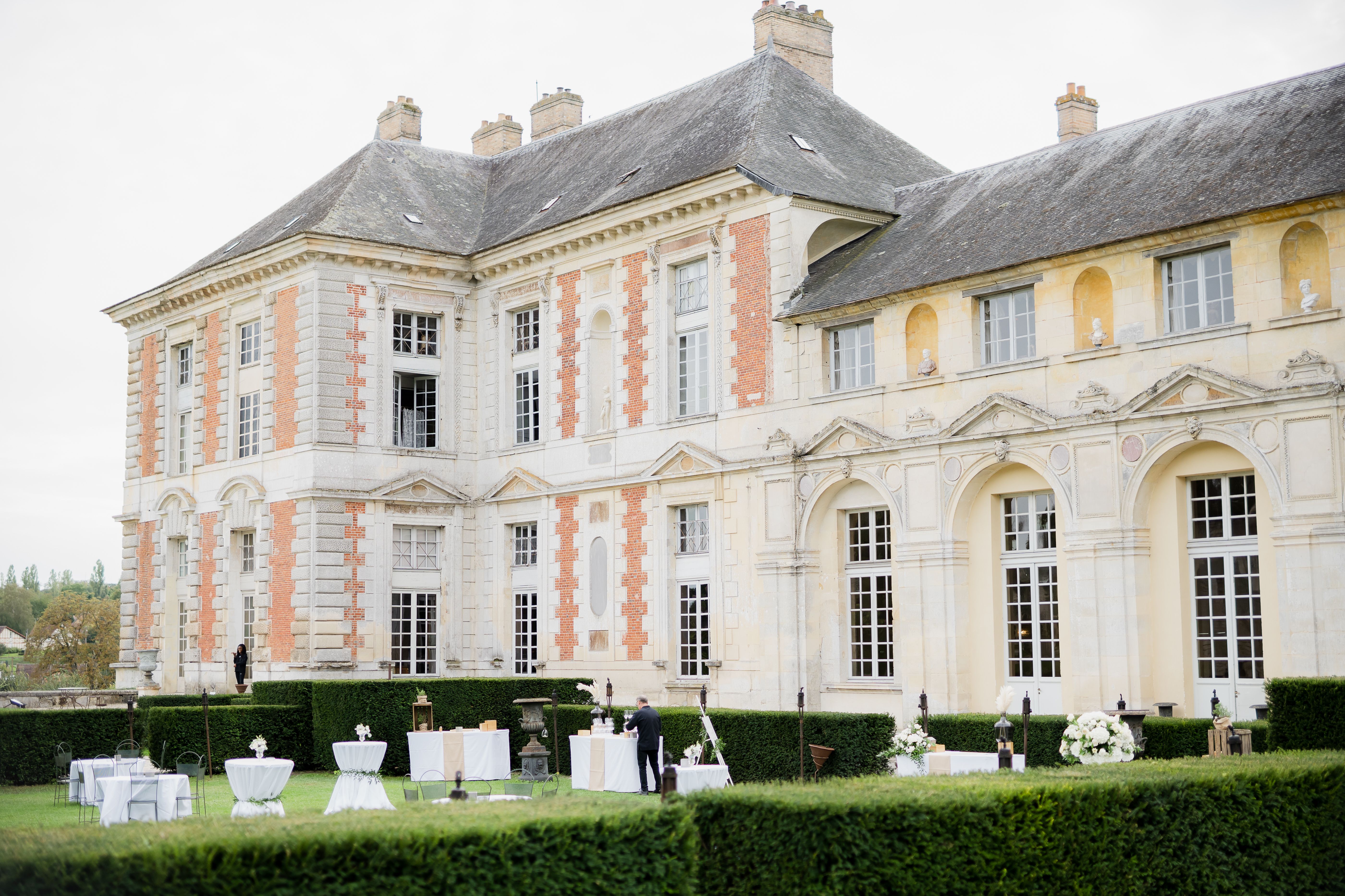 A wide shot of the rear facade of a large French chateau featuring cream stone and terracotta brick detailing, mansard roofing, and classical architectural ornamentation including arched windows and sculpted pediments. The chateau grounds are being set up for an outdoor cocktail hour, with white cocktail tables draped in white linen, seated reception tables with white chair covers, and lanterns placed along the terrace. White floral arrangements, including a large white bloom cluster visible on the right, are positioned throughout the setup. One staff member in a dark suit is visible attending to the bar area, and a second person stands near the chateau entrance. The decor palette is white and ivory with a classic, formal styling theme. Potential venue feature image.