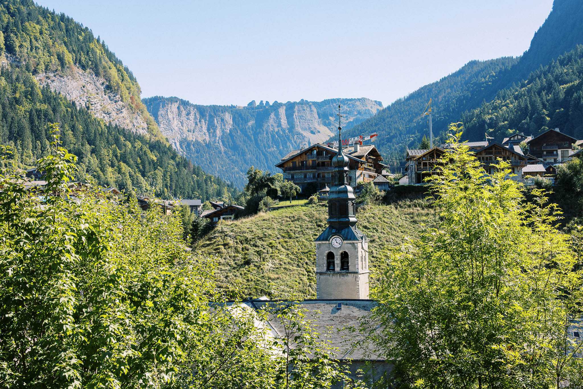 Wide-shot exterior view of a French Alpine village featuring a church bell tower with a distinctive dark metal onion-shaped spire and clock face at its center, surrounded by traditional wood-and-stone chalet-style buildings. No people, wedding party, or wedding-specific decor are visible in this image. The shot frames the church tower and village architecture against a backdrop of forested mountain ridges. Potential venue feature image.