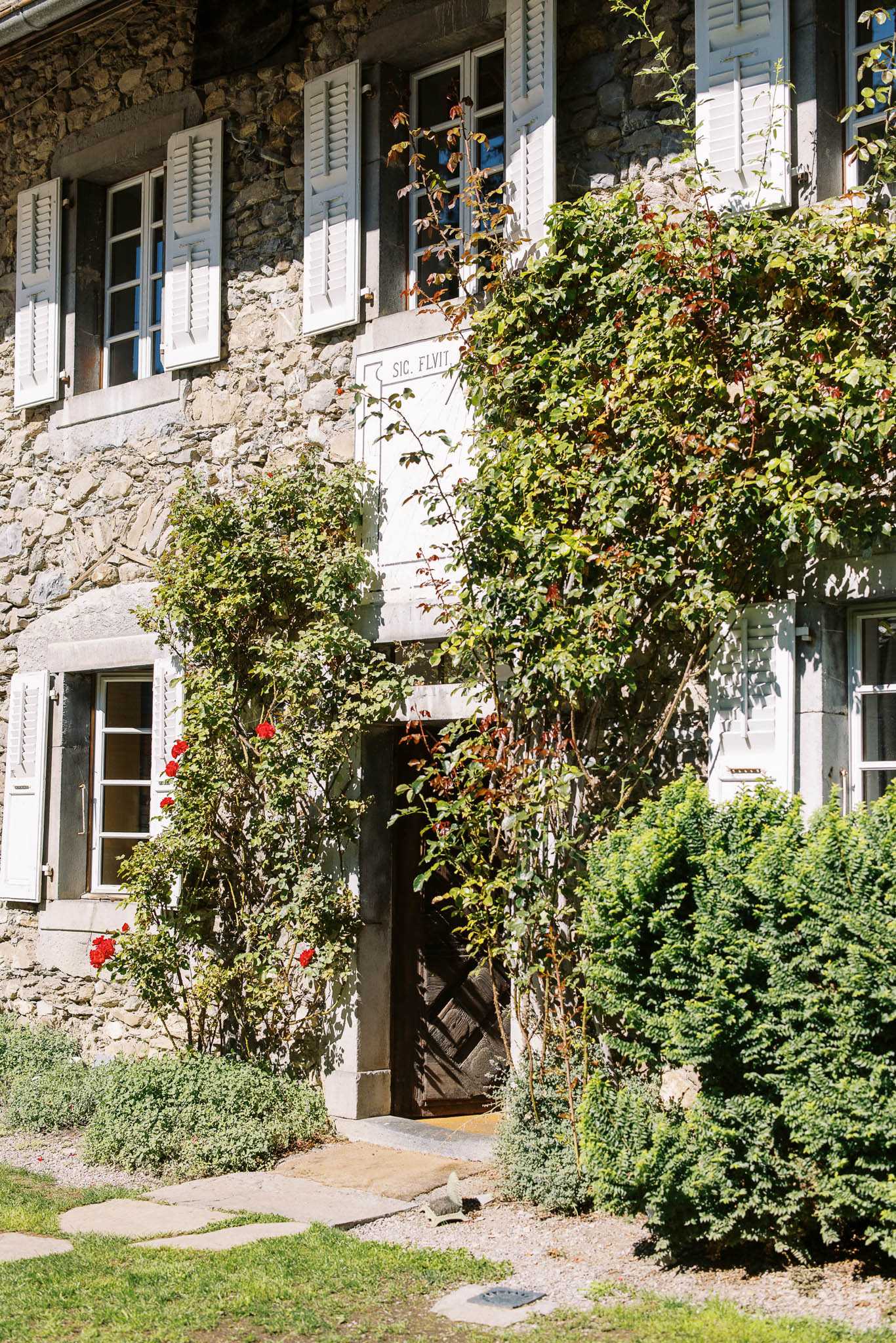 Exterior architectural shot of a historic stone manor house, showing a two-story facade with white-painted shutters on multiple windows and a heavy wooden arched entrance door. Climbing roses with red blooms and dense green vines trail up the stone walls on either side of the doorway, and a carved stone plaque above the entrance reads 'SIC. FLVIT'. The foreground includes a stone pathway, trimmed shrubs, and a lawn. No people are visible in the frame. Wide shot, vertical composition, shot in full color with bright natural daylight. Potential venue feature image.