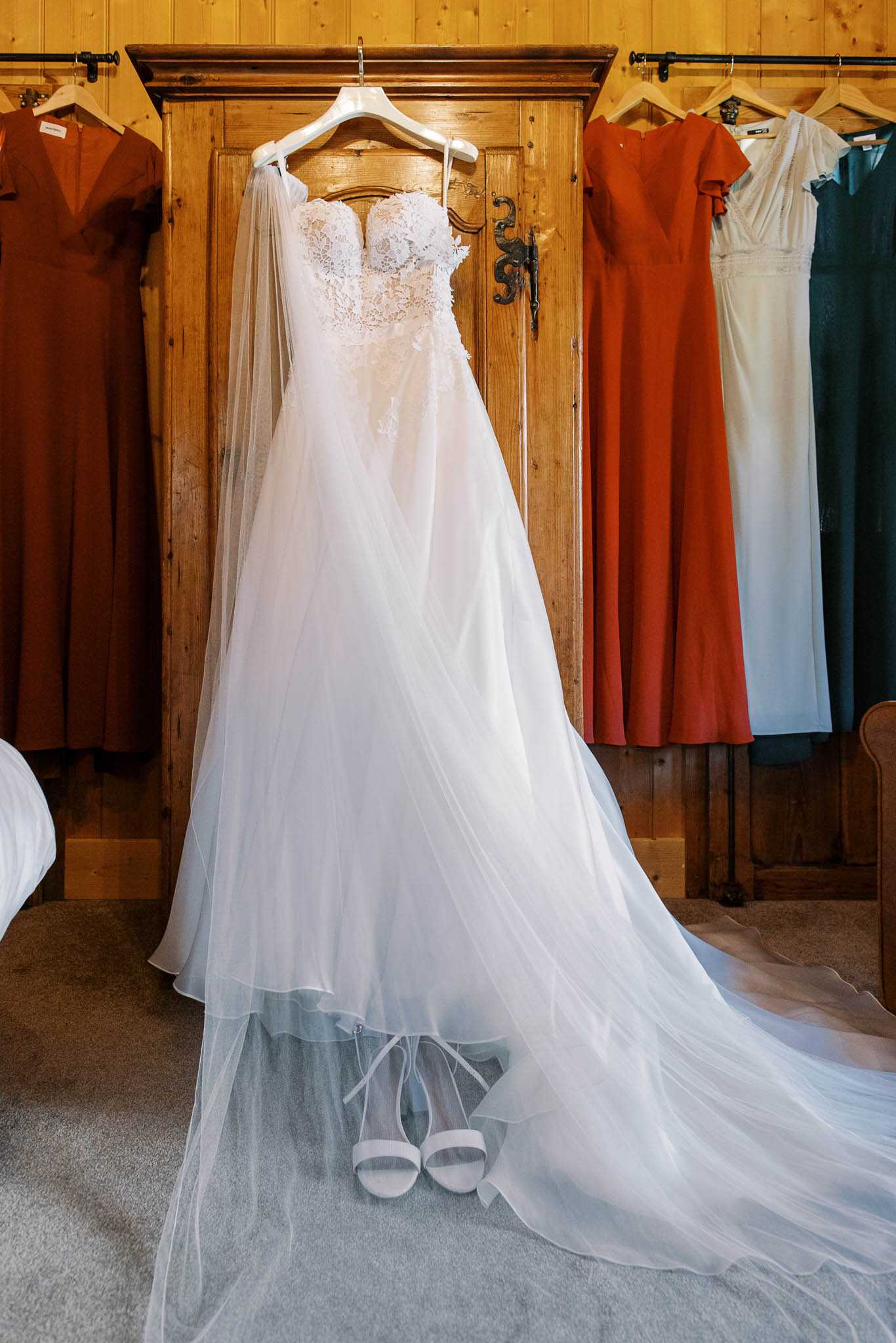 A getting-ready flat-lay style detail shot taken indoors in what appears to be a bridal suite with warm pine wood paneling and a large wooden armoire. The bride's ivory strapless wedding gown is hung on a white hanger against the armoire door, featuring a lace appliqué bodice and a full, flowing tulle skirt with a long train that pools on the floor. A pair of white strappy block-heel sandals is placed beneath the dress on the carpeted floor. Flanking the armoire on both sides are bridesmaid dresses hanging on wooden hangers — the majority are terracotta/burnt orange in color with varying necklines, alongside one ivory dress and one teal dress, suggesting a mix-and-match bridesmaid styling approach. The composition is a full-length portrait shot centered on the bridal gown.