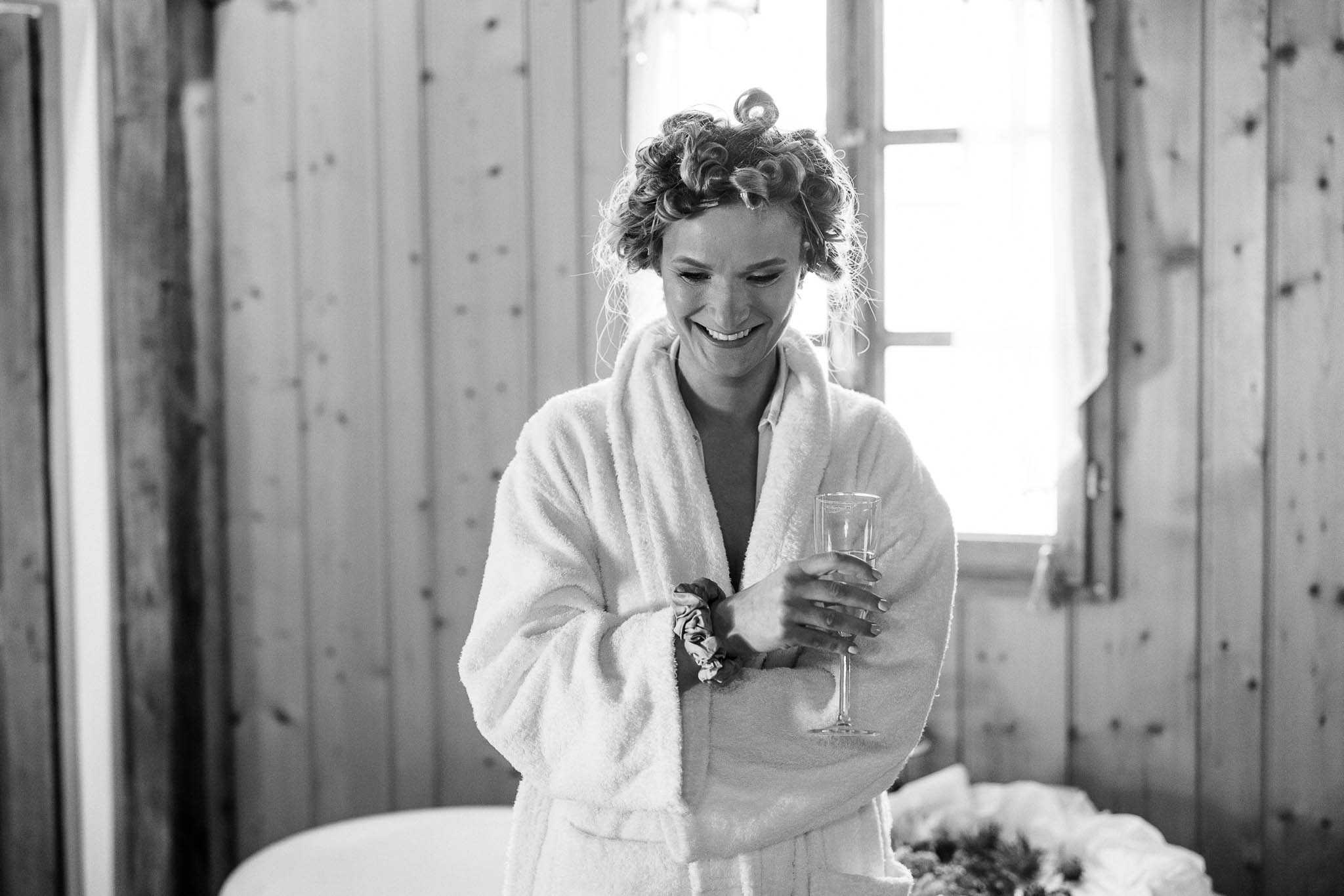 A black-and-white getting-ready shot of a woman, likely the bride, laughing and looking downward while holding a champagne flute. She is wearing a fluffy white bathrobe with hair curlers in her curly hair and a scrunchie on her wrist. The setting is an indoor room with wood-paneled walls and a small window with sheer curtains letting in bright natural light from behind. The composition is a mid-length portrait with soft contrast, and the bouquet is partially visible in the lower right corner of the frame.