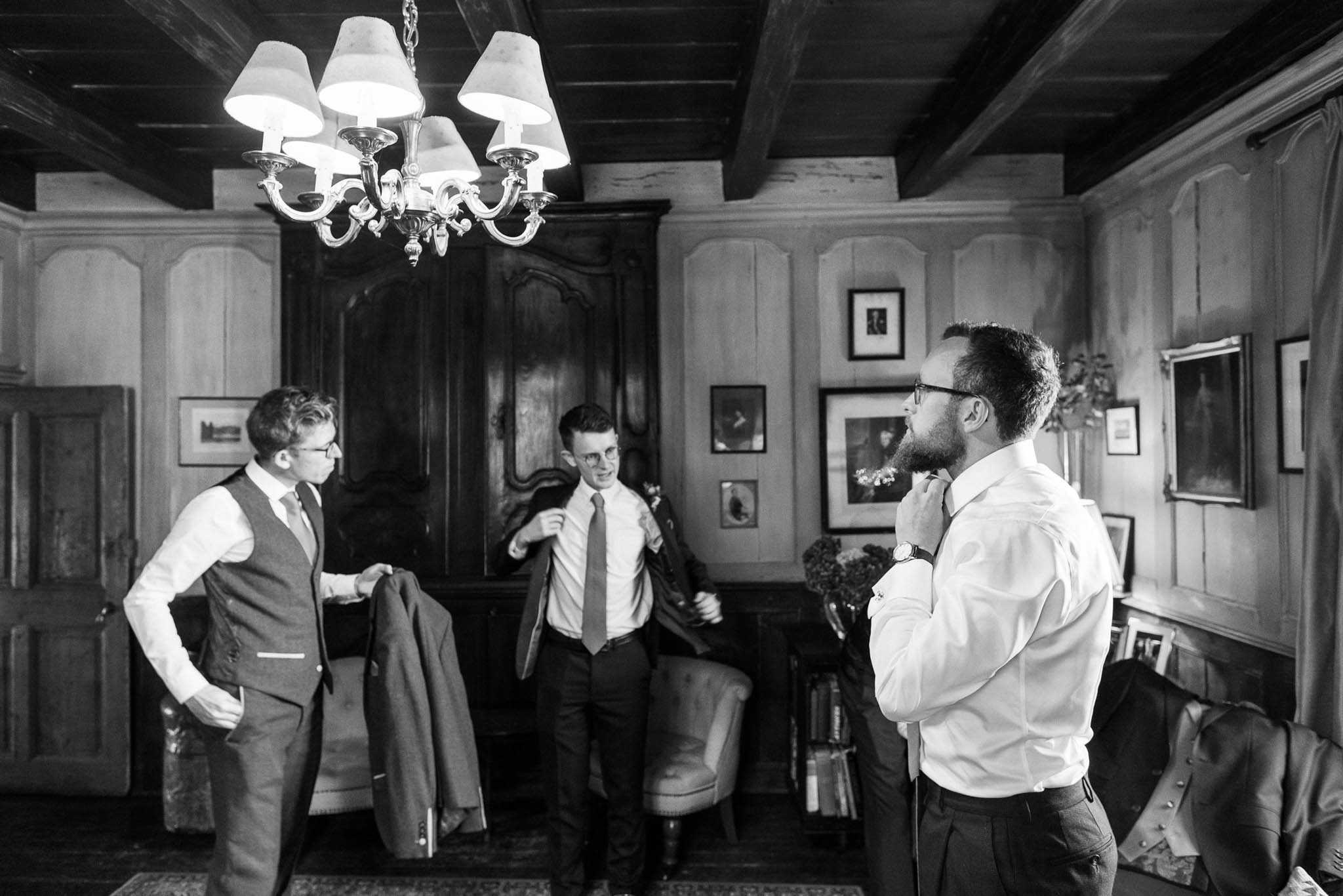 A black-and-white getting-ready shot of three men in a wood-paneled interior room, likely inside a chateau or historic manor. The man on the right, bearded and wearing glasses, adjusts his collar or tie while still in his dress shirt, with a suit jacket visible on a chair beside him; he wears a watch on his left wrist. In the center, a second man in a tie and dress shirt is putting on or adjusting his suit jacket, and to the left, a third man wearing a waistcoat and tie holds a suit jacket over his arm. The room features dark exposed ceiling beams, a multi-arm chandelier with lampshades, a large dark wood armoire, upholstered armchairs, and walls hung with framed artwork, giving the space a classic, old-house character. The image is a medium wide shot with moderate contrast, capturing a candid, relaxed moment between the men as they dress before the ceremony.
