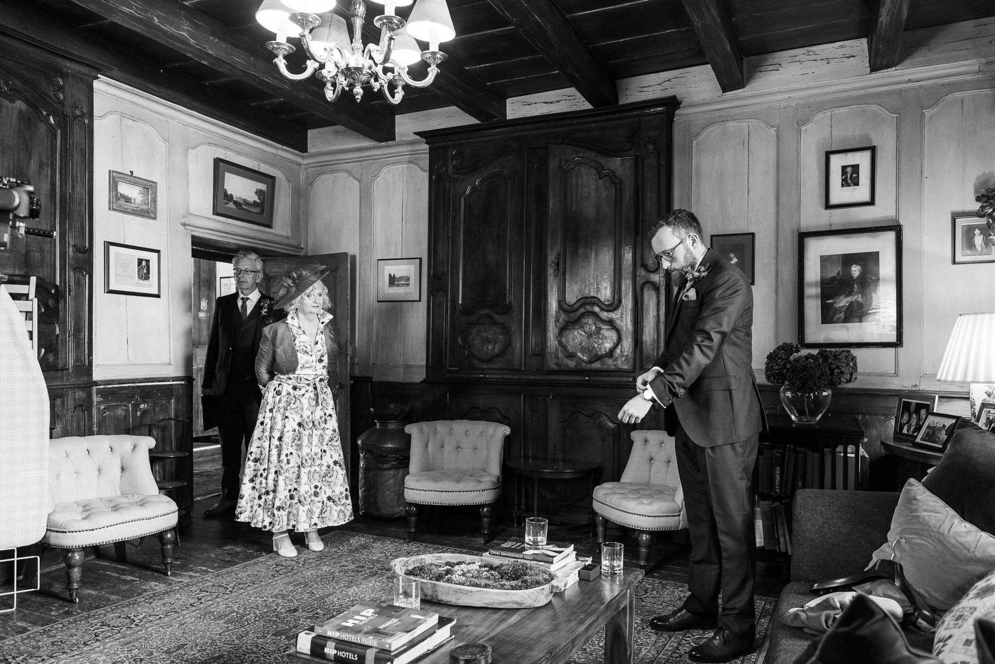 Black-and-white wide shot taken indoors in a richly furnished salon or drawing room, likely within a historic chateau or manor house, featuring dark exposed ceiling beams, wood-paneled walls, a crystal chandelier, antique armchairs, and a large carved wooden armoire. The groom, wearing a dark suit with a boutonniere and glasses, stands to the right adjusting his cufflinks or watch, while two guests — a man in a dark suit with a lapel flower and a woman in a floral midi dress and wide-brimmed hat — stand in the background near a doorway. The room is decorated with framed portraits and landscape paintings on the walls, a coffee table with stacked books and glassware in the foreground, and a vase of flowers on a side table. The scene appears to be a pre-ceremony getting-ready or gathering moment, captured in a candid, documentary style with strong mid-tone contrast.