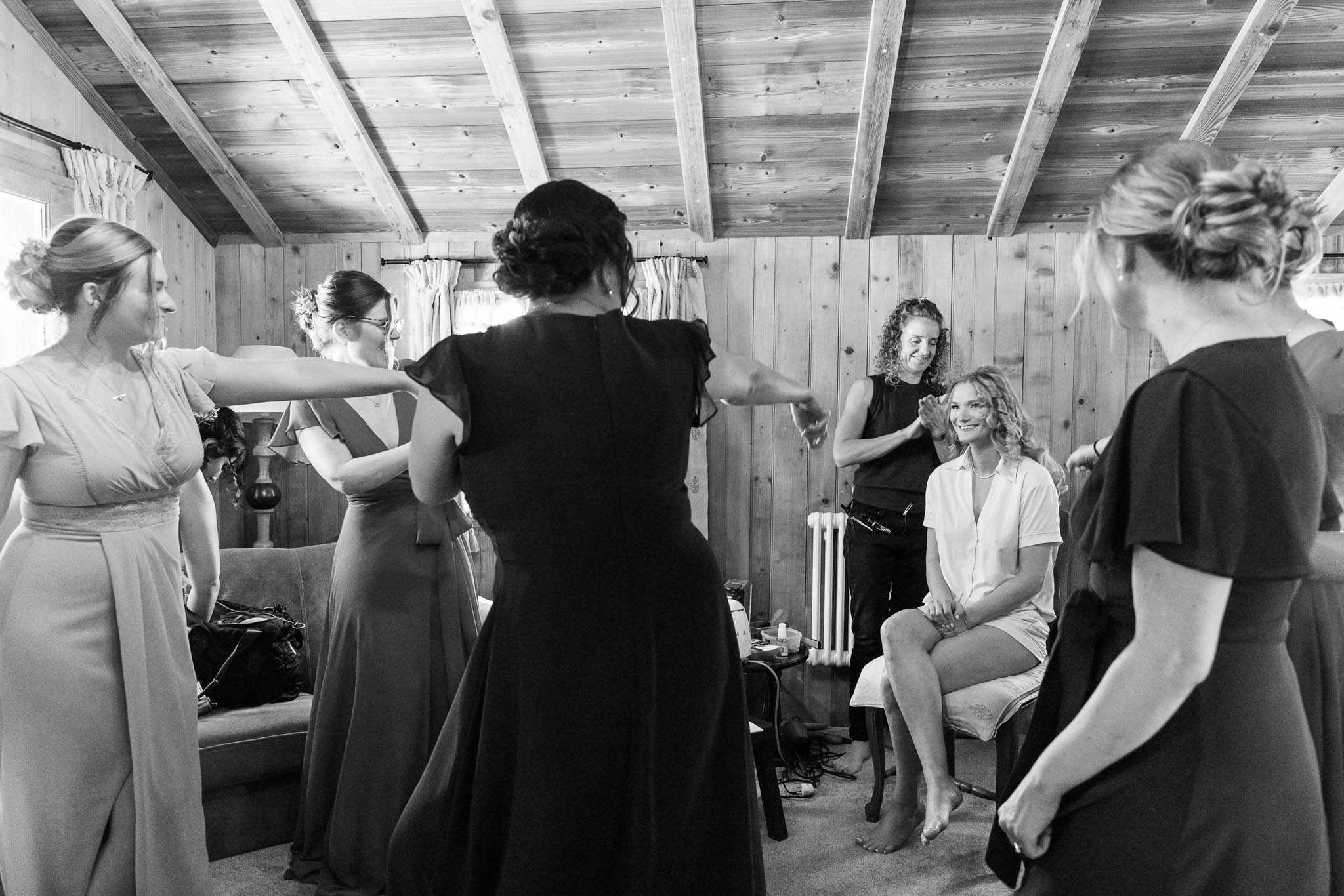 A black-and-white getting-ready scene captured indoors in a wood-paneled room with exposed timber ceiling beams, likely a cabin or rustic lodge. The bride, dressed in a casual white top and shorts, sits barefoot in a chair smiling broadly while a hair or makeup artist works on her; a small side table with beauty products is visible nearby. Several bridesmaids in floor-length dresses with upswept hairstyles gather around her, with two appearing to playfully reach out or gesture toward each other in a candid, lively moment. The image is a wide candid shot with strong contrast between the dark clothing of the stylists and the lighter tones of the bridesmaids' gowns and wood-paneled walls.