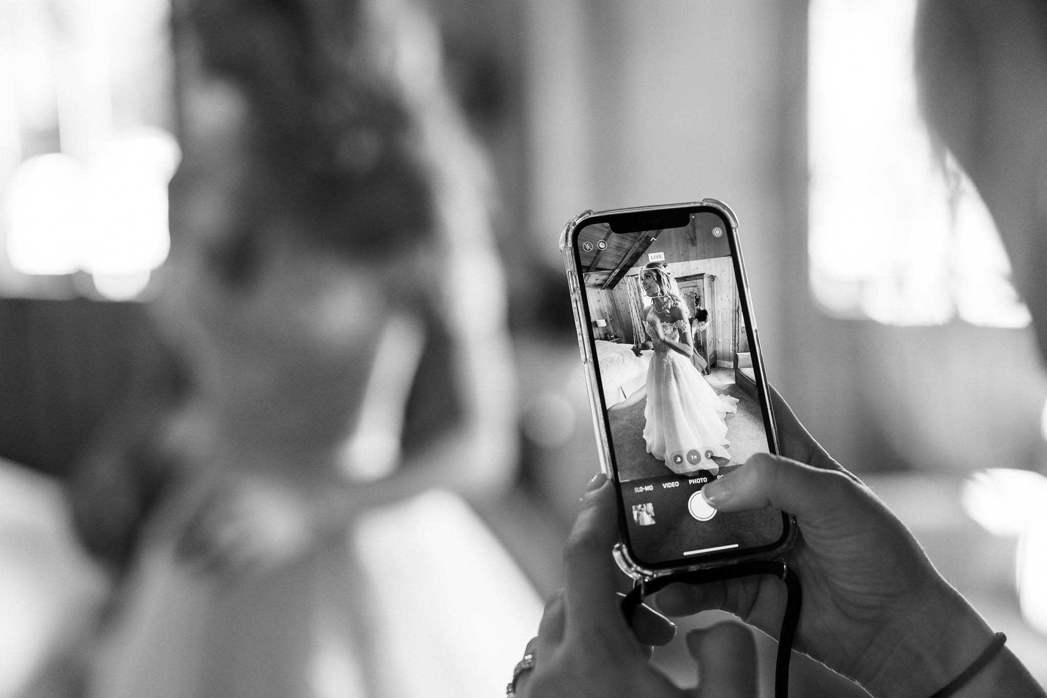 This black-and-white close-up shot captures a getting-ready moment indoors, where a guest or bridesmaid holds an iPhone in a clear case to photograph the bride. The phone screen is sharp and in focus, showing the bride in full in a full-skirted white wedding gown with an embellished bodice, standing in a rustic wood-beamed room in front of what appears to be an antique armoire or doorway. The bride herself is blurred in the background on the left side of the frame, with the back of her styled updo and the top of her dress visible. The composition uses a shallow depth of field to draw focus entirely to the phone screen, creating a layered, candid feel with strong contrast between the bright highlights from the windows and the darker mid-tones.