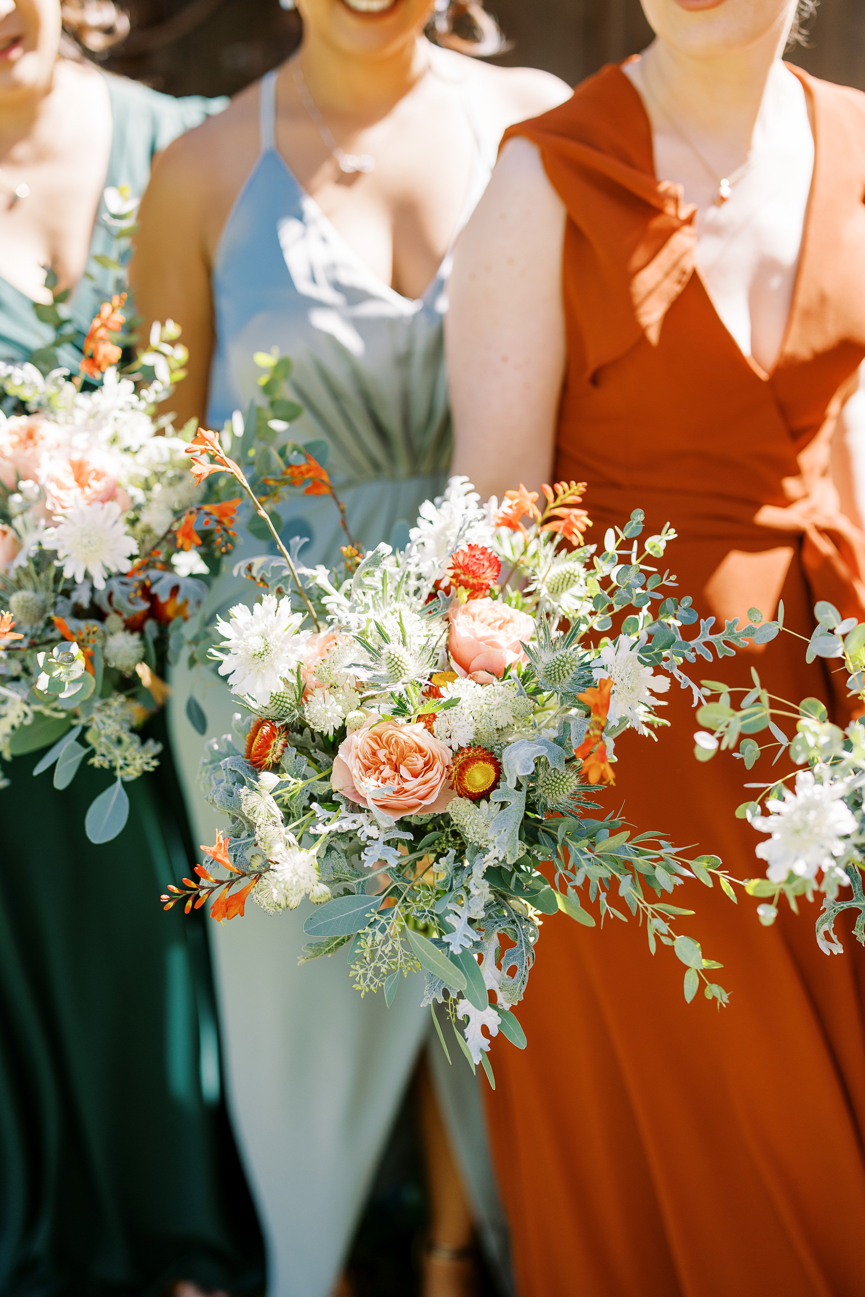 A close-up portrait of three bridesmaids holding bouquets, photographed from the neck down in bright natural light. The bridesmaids are wearing mismatched dresses in forest green, sage/mist blue, and burnt orange. The bouquets are loosely arranged in a garden-style with trailing eucalyptus, white scabiosa, blush garden roses, white chrysanthemums, blue-green thistles, orange crocosmia, and straw flowers in rust and yellow tones. The overall floral palette combines peach, white, burnt orange, and sage green with an organic, unstructured composition. The image is a mid-shot detail focusing on the dresses and florals rather than the bridesmaids' faces.