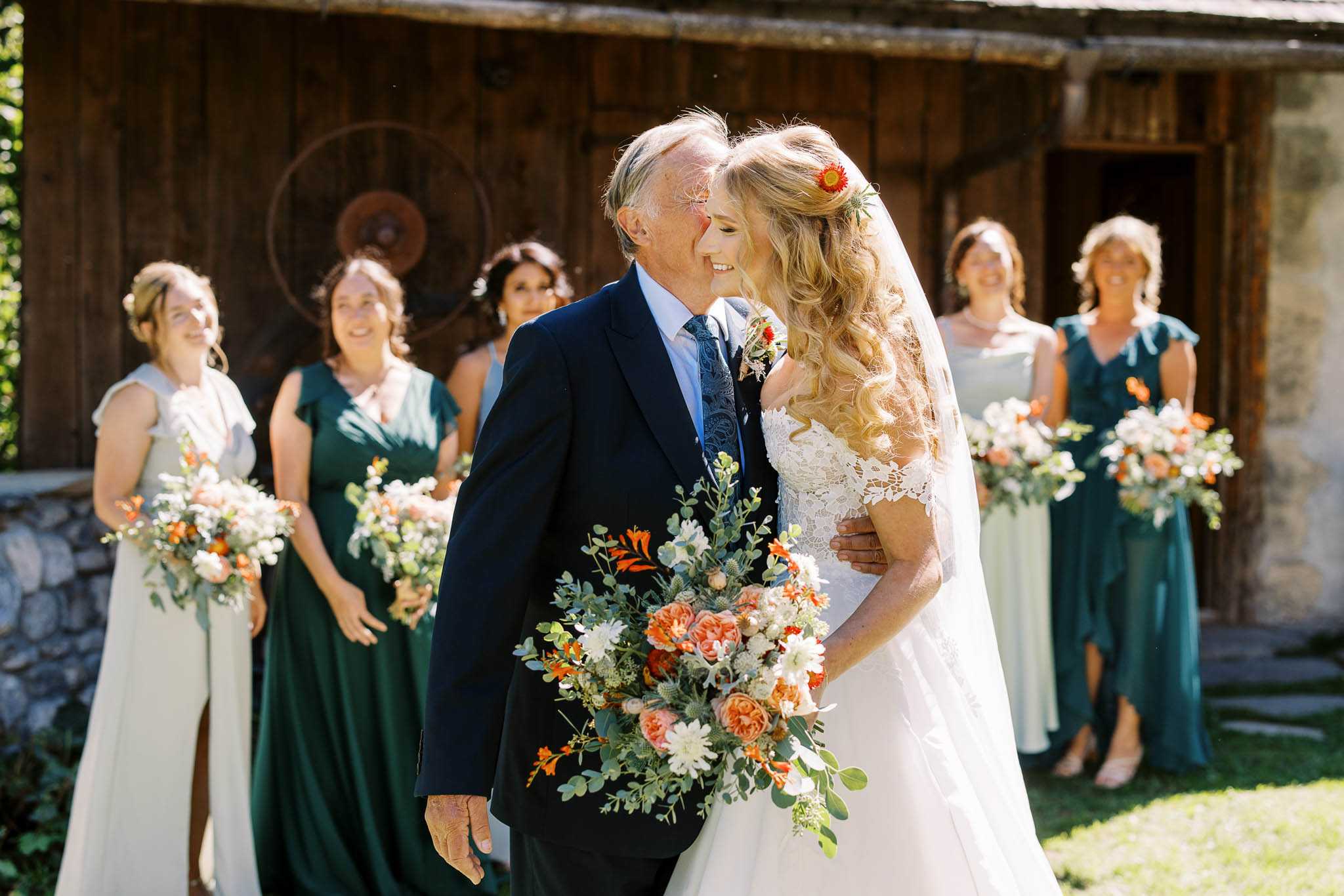 An outdoor bridal party portrait taken before the ceremony, showing a bride sharing a close, smiling moment with an older man — likely her father — who is dressed in a navy suit with a blue patterned tie and a boutonniere. The bride wears a lace off-the-shoulder wedding dress with a veil, her blonde wavy hair half-up with a small orange gerbera daisy hair accessory. She holds a large loose bouquet of coral garden roses, orange gerberas, white daisy-like blooms, thistles, and eucalyptus foliage. Behind them, six bridesmaids stand in a mix of deep forest green and pale sage/ivory floor-length dresses, each holding smaller versions of the same orange and white floral arrangements. The setting is outdoor in bright sunlight in front of a rustic weathered wooden barn structure, giving the scene a boho-rustic aesthetic. The shot is a medium portrait with the bridal party softly out of focus in the background.