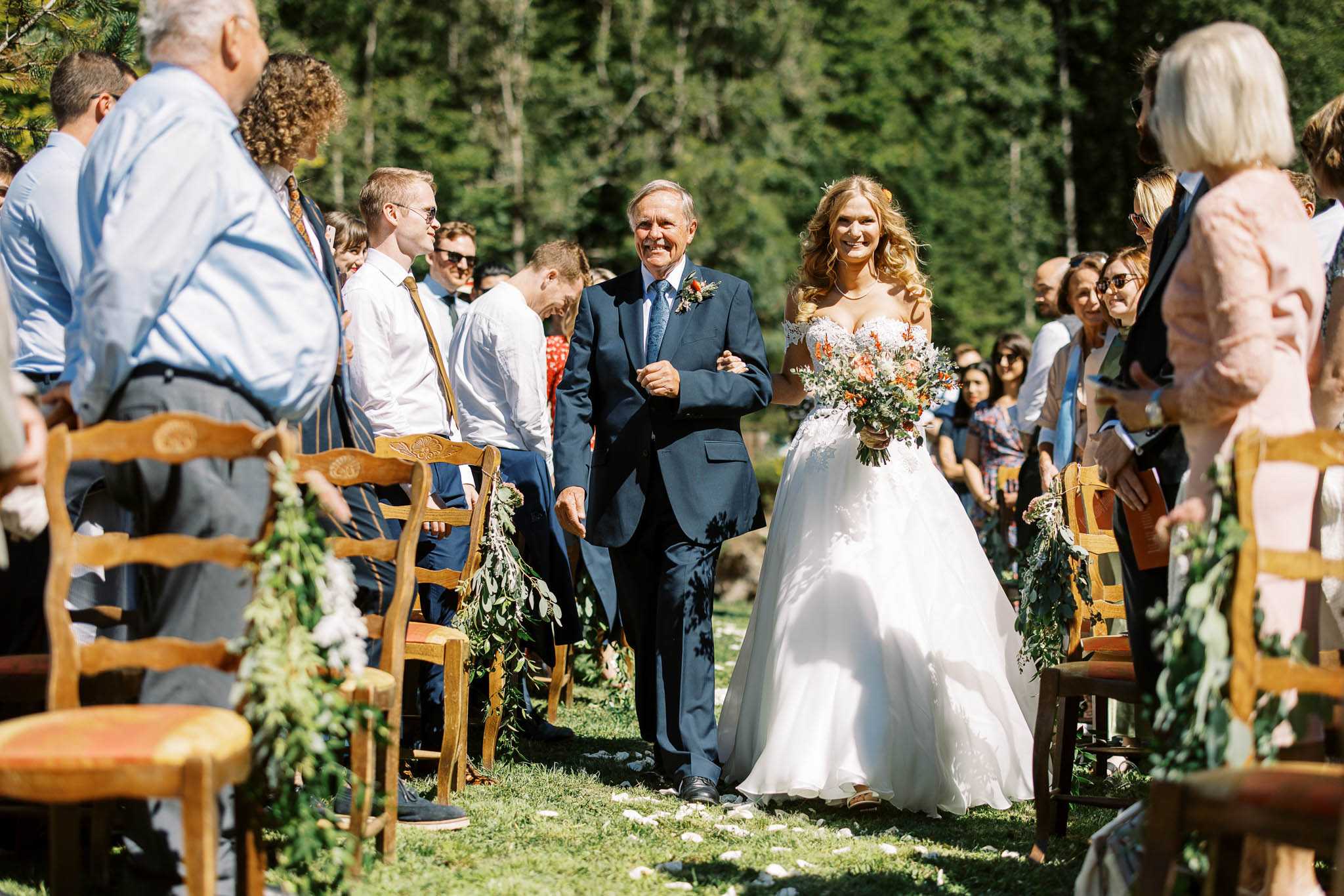 The bride is being walked down the aisle by an older man, likely her father, during an outdoor ceremony set on a grass lawn surrounded by tall conifer trees. The bride wears a white ballgown with lace appliqué detailing and a sweetheart neckline, and carries a loose, garden-style bouquet featuring burnt orange, terracotta, and white blooms with trailing greenery. The escort wears a navy suit with a teal-blue tie and a boutonnière with orange accents. Wooden farmhouse-style chairs line the aisle, decorated with cascading greenery garlands, and white flower petals are scattered on the grass. A crowd of approximately 40–50 standing guests looks on from both sides of the aisle. The shot is a wide, eye-level perspective taken from within the seated guest area looking toward the couple.