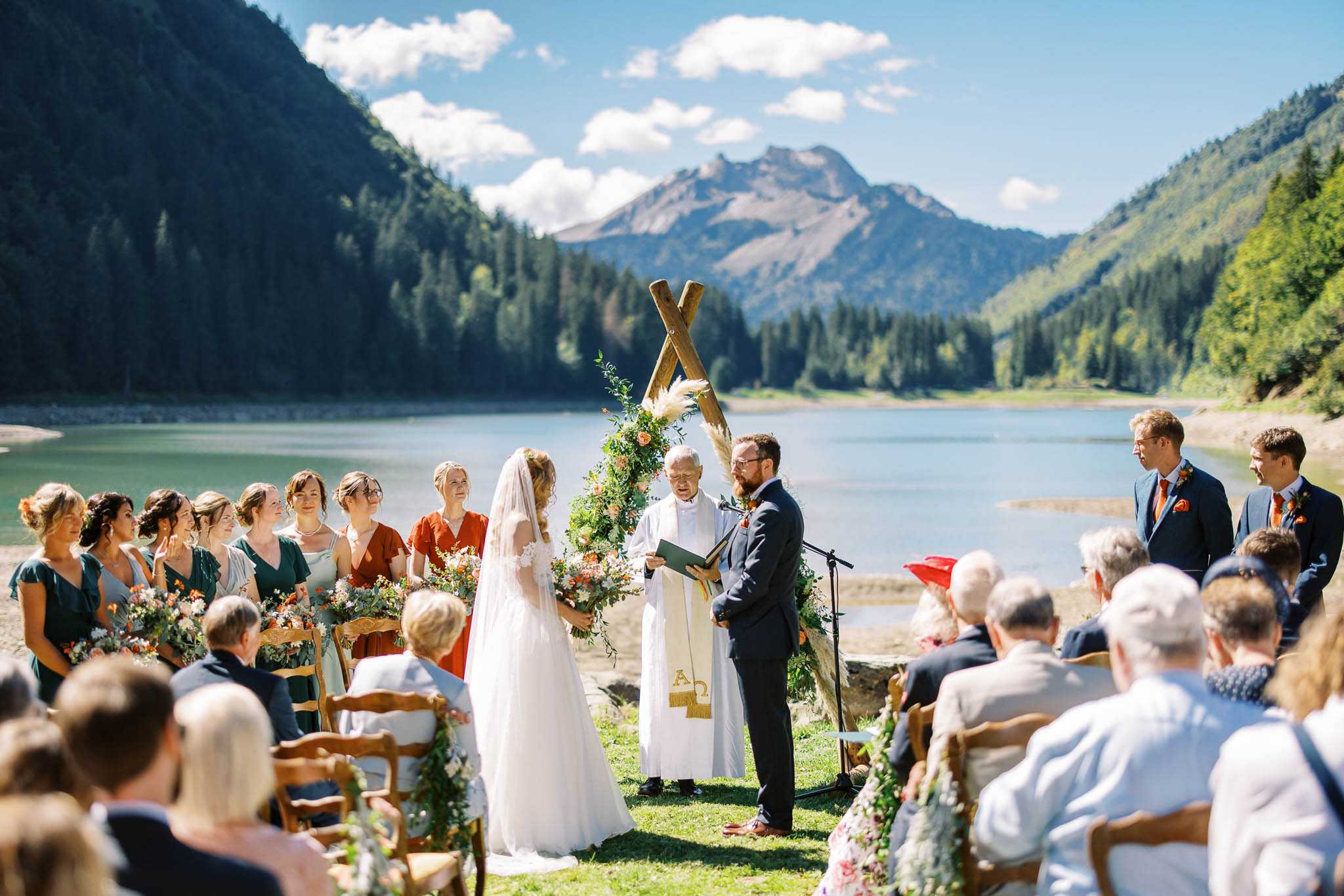 An outdoor wedding ceremony taking place on the grassy shore of a mountain lake, with a dramatic alpine backdrop. The bride wears a white gown with a long veil and holds a bouquet of coral, orange, and greenery blooms, while the groom wears a navy suit with an orange boutonniere; an officiant in white robes with a gold-fringed stole reads from a book beneath a rustic crossed-log arch decorated with pampas grass, coral and peach blooms, and trailing greenery. The bridal party of approximately eight bridesmaids wear forest green and rust/terracotta wrap dresses and carry wildflower bouquets in warm orange and coral tones, while two groomsmen in navy suits with orange ties stand on the opposite side. Seated guests of approximately 30 people on wooden chairs face the altar, with the aisle edges lined with floral arrangements; the overall styling is bohemian-rustic with a warm autumnal color palette. Wide shot taken from slightly behind the seated guests.
