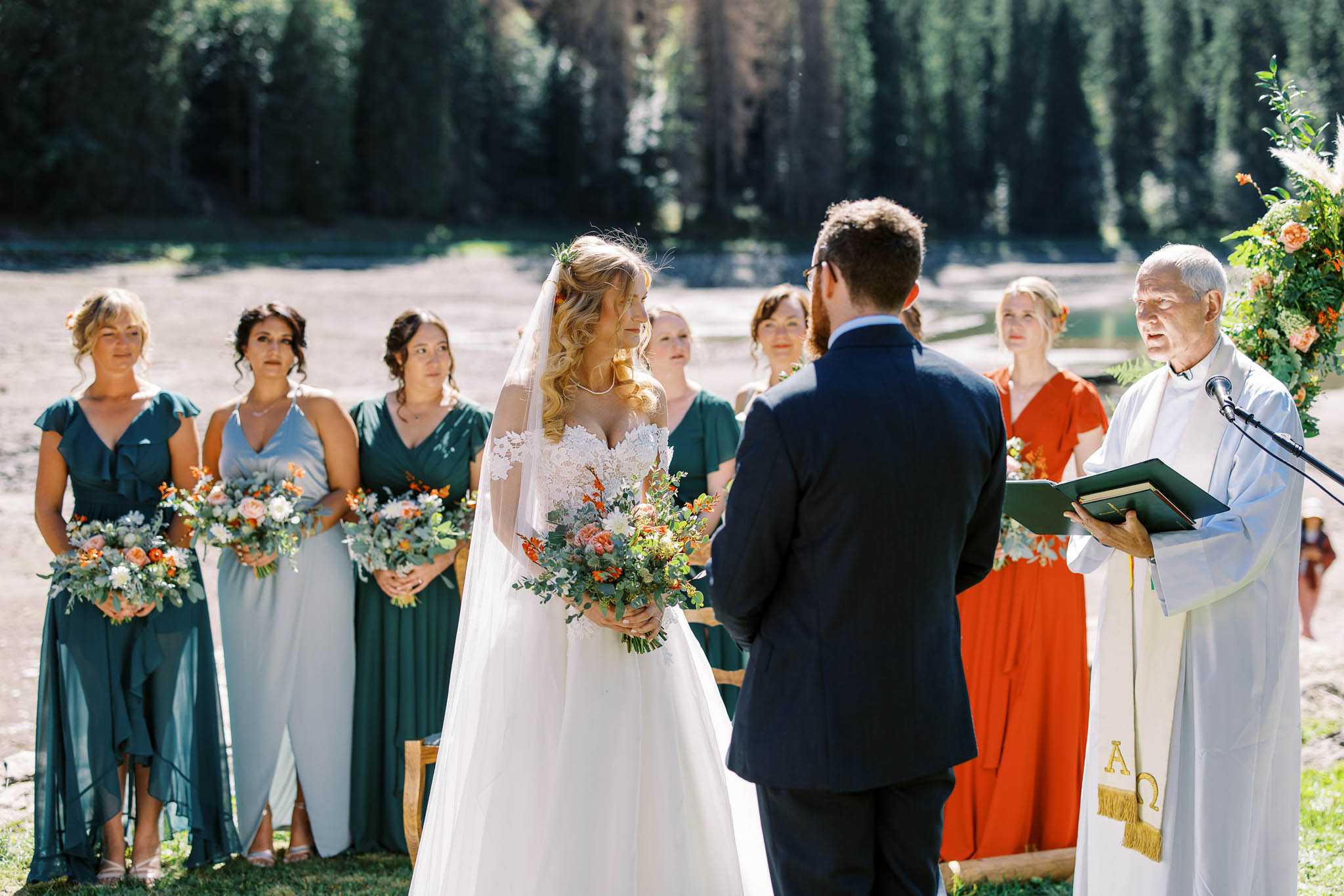An outdoor wedding ceremony taking place in a natural setting, with the couple standing at the altar facing each other while an officiant in white robes and a gold Alpha-Omega stole reads from a green book at a microphone stand. The bride wears an off-the-shoulder white ballgown with lace bodice detailing and a long veil, holding a loose bouquet of peach roses, orange blooms, white flowers, and eucalyptus. The groom wears a navy suit. Four bridesmaids stand to the left in a mix of teal and sage green dresses, each holding matching bouquets with orange and peach flowers and greenery. A guest in a rust-orange dress stands in the background alongside other seated guests. A circular floral arch partially visible on the right features orange blooms and lush greenery. The image is a medium wide shot taken in bright natural daylight.