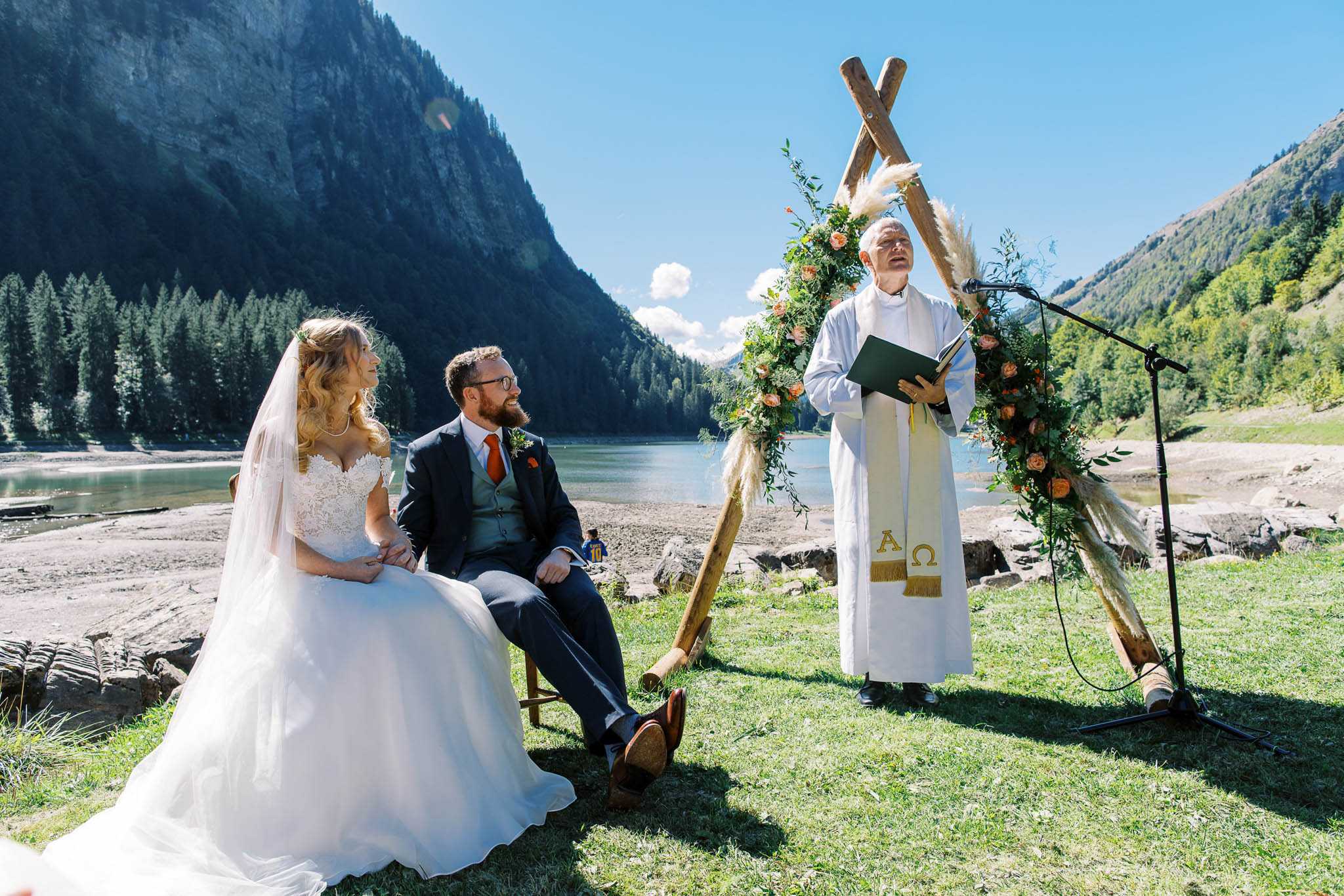 An outdoor wedding ceremony taking place on the grassy bank of a mountain lake, with the couple seated in chairs facing a officiant who is reading aloud from a dark green book. The bride wears a white lace off-the-shoulder ballgown with a cathedral-length veil, and the groom wears a charcoal suit with a grey waistcoat and burnt orange tie, with a matching orange buttonhole. The officiant is dressed in white robes with a gold-embroidered stole bearing Alpha and Omega symbols. Behind them stands a rustic wooden A-frame arch decorated with peach/coral roses, pampas grass, and trailing greenery. The wide-angle shot captures the full lakeside setting with mountain peaks and conifer forests in the background, giving the ceremony a boho-rustic alpine aesthetic.