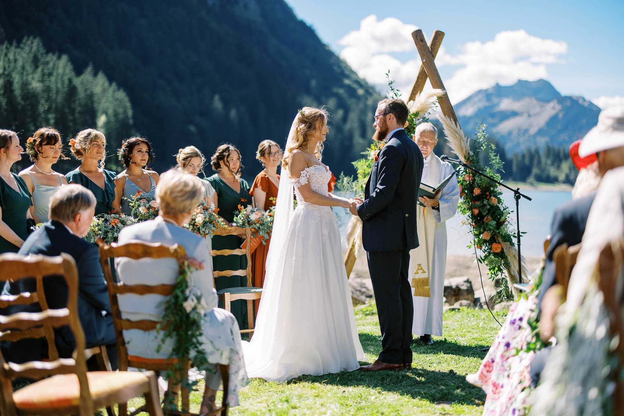 An outdoor wedding ceremony taking place on a grassy lakeside setting with mountain peaks visible in the background. The bride, wearing an off-the-shoulder white lace ballgown with a veil, and the groom, in a dark navy suit, stand facing each other holding hands before an officiant in white robes, who reads from a book at a microphone stand. Behind the couple stands a rustic wooden X-shaped arch decorated with trailing greenery, pampas grass, and peach and orange floral clusters. Approximately six bridesmaids are lined up to the left, wearing a mix of forest green and terracotta/rust-colored dresses, holding bouquets of peach roses, orange blooms, and eucalyptus. Seated guests on wooden chairs are visible in the foreground on both sides of the aisle. The overall styling is rustic-boho with an earthy color palette of forest green, terracotta, and peach. Wide mid-shot capturing the full ceremony scene.