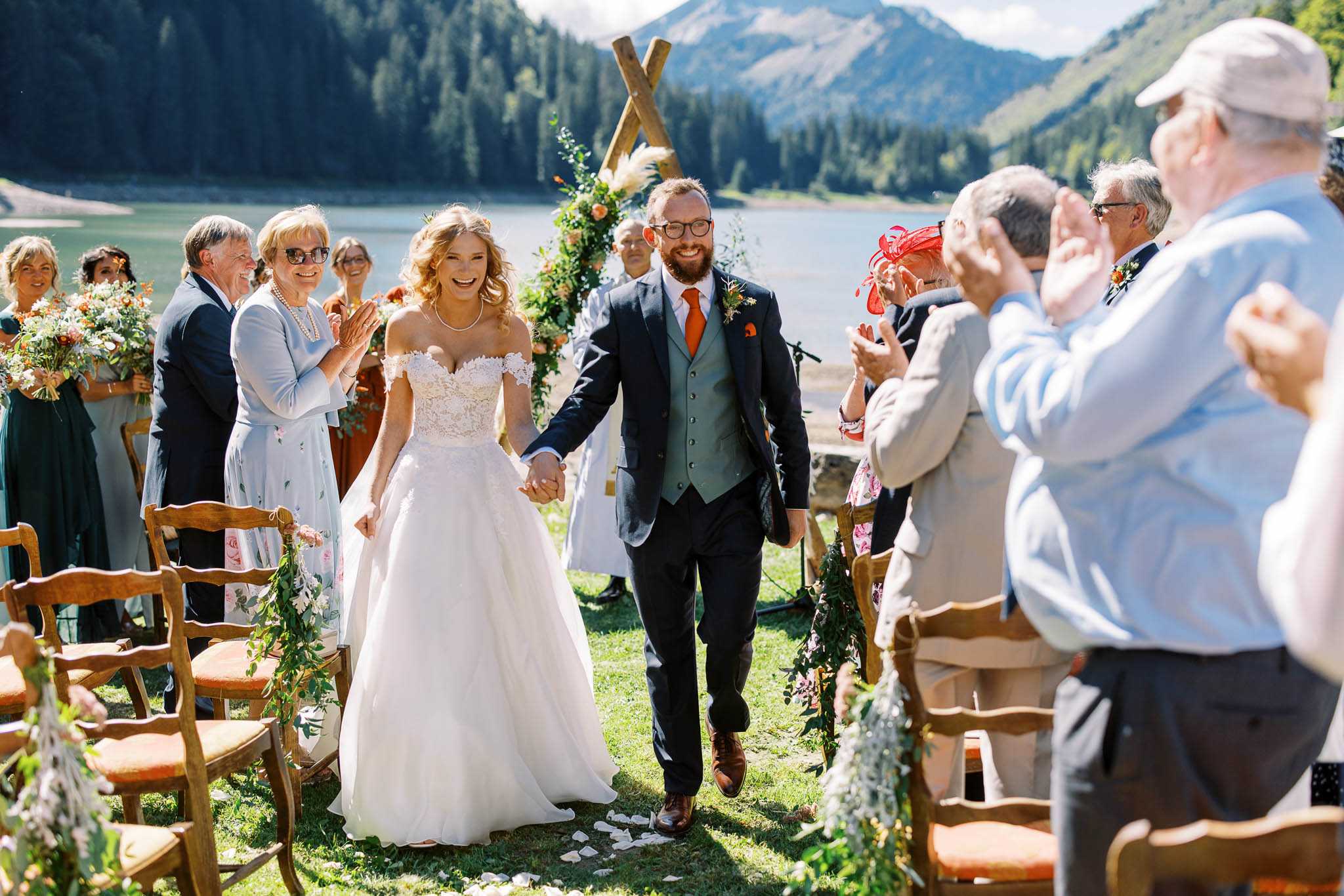 The bride and groom walk back up the aisle together following an outdoor ceremony, holding hands and laughing, surrounded by approximately 20 applauding guests. The setting is a lakeside mountain location with a rustic wooden X-shaped arch behind them decorated with pampas grass and orange and peach florals. The bride wears an off-the-shoulder ivory lace ballgown with a full skirt and a pearl necklace, while the groom wears a navy suit with a grey waistcoat, burnt orange tie, and brown leather shoes. Wooden chairs with orange cushions line the aisle, decorated with greenery and white florals. Bridesmaids in teal/dark green dresses are visible on the left holding bouquets of orange, rust, and white blooms. The overall decor palette combines earthy autumn tones — burnt orange, rust, and greenery — with a rustic alpine style. This is a wide mid-shot taken at ground level capturing the recessional moment.