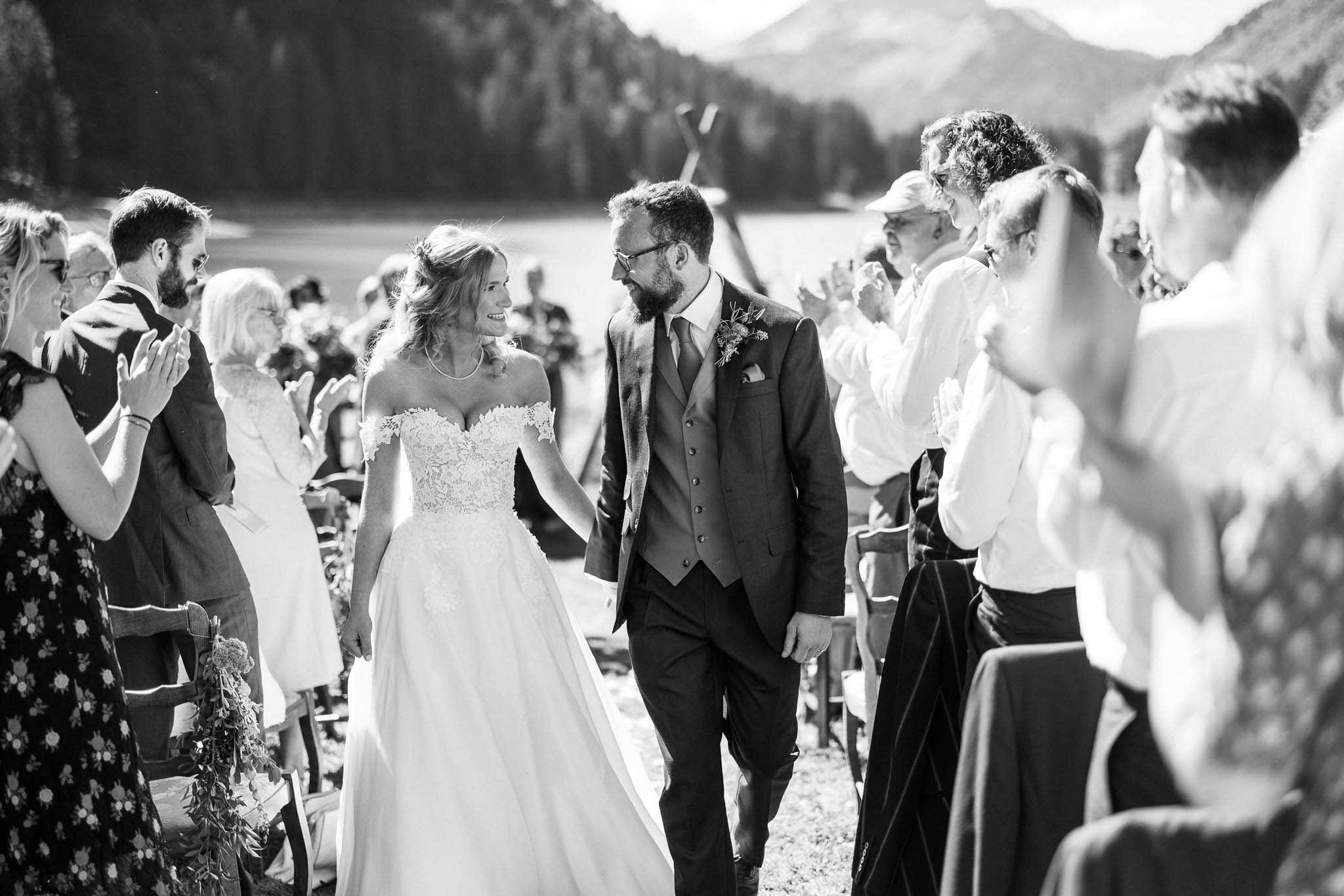 This black-and-white image captures the couple's recessional during an outdoor ceremony, with the bride and groom walking arm-in-arm down the aisle while approximately 20–25 guests lining both sides applaud. The bride wears an off-the-shoulder lace bodice ballgown with a flowing skirt, accessorized with a delicate necklace and soft updo with loose waves, while the groom wears a three-piece suit with a tie and a small floral boutonniere. The ceremony appears to take place on open ground with wooden chairs and aisle décor that includes a trailing greenery arrangement visible on one chair. The image is a mid-range shot with strong contrast and bright highlights, and the couple are gazing at each other and smiling.