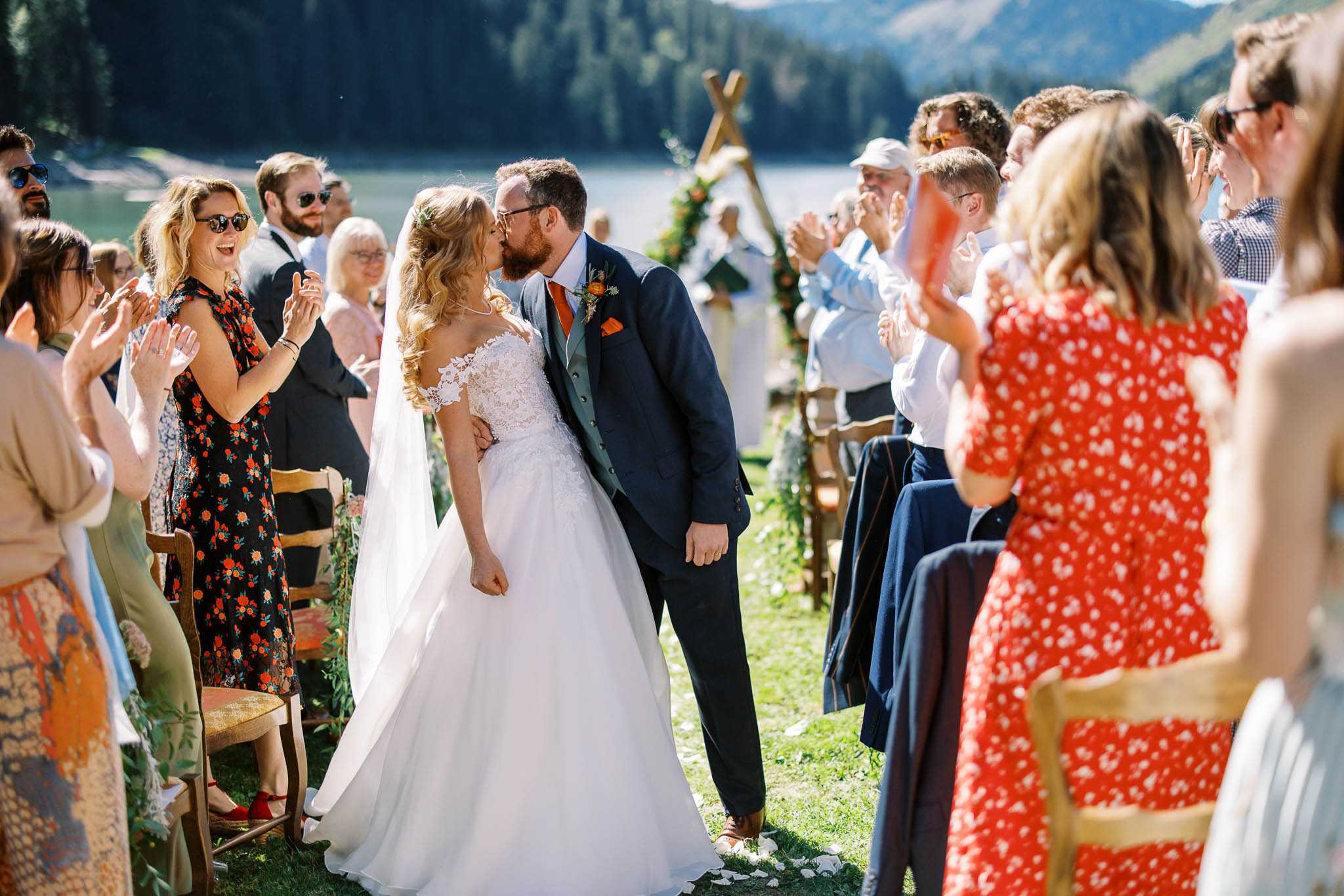 The bride and groom share their first kiss at the end of the aisle during an outdoor ceremony, with approximately 30 guests on either side applauding and cheering. The setting is a lakeside ceremony on a grass lawn, with a rustic wooden triangular arch decorated with foliage and orange and warm-toned florals visible in the background. The bride wears an off-the-shoulder ball gown with lace bodice and a long veil, while the groom wears a navy suit with a sage green waistcoat, an orange tie, and a burnt orange boutonnière. Guests are seated and standing on wooden chairs, dressed in a mix of casual summer attire including floral print dresses in red and black. The decor palette centers around burnt orange and warm earthy tones in a relaxed, boho-rustic style. The image is a mid-range portrait shot taken in bright natural sunlight with a shallow depth of field that blurs the background guests and mountain scenery.