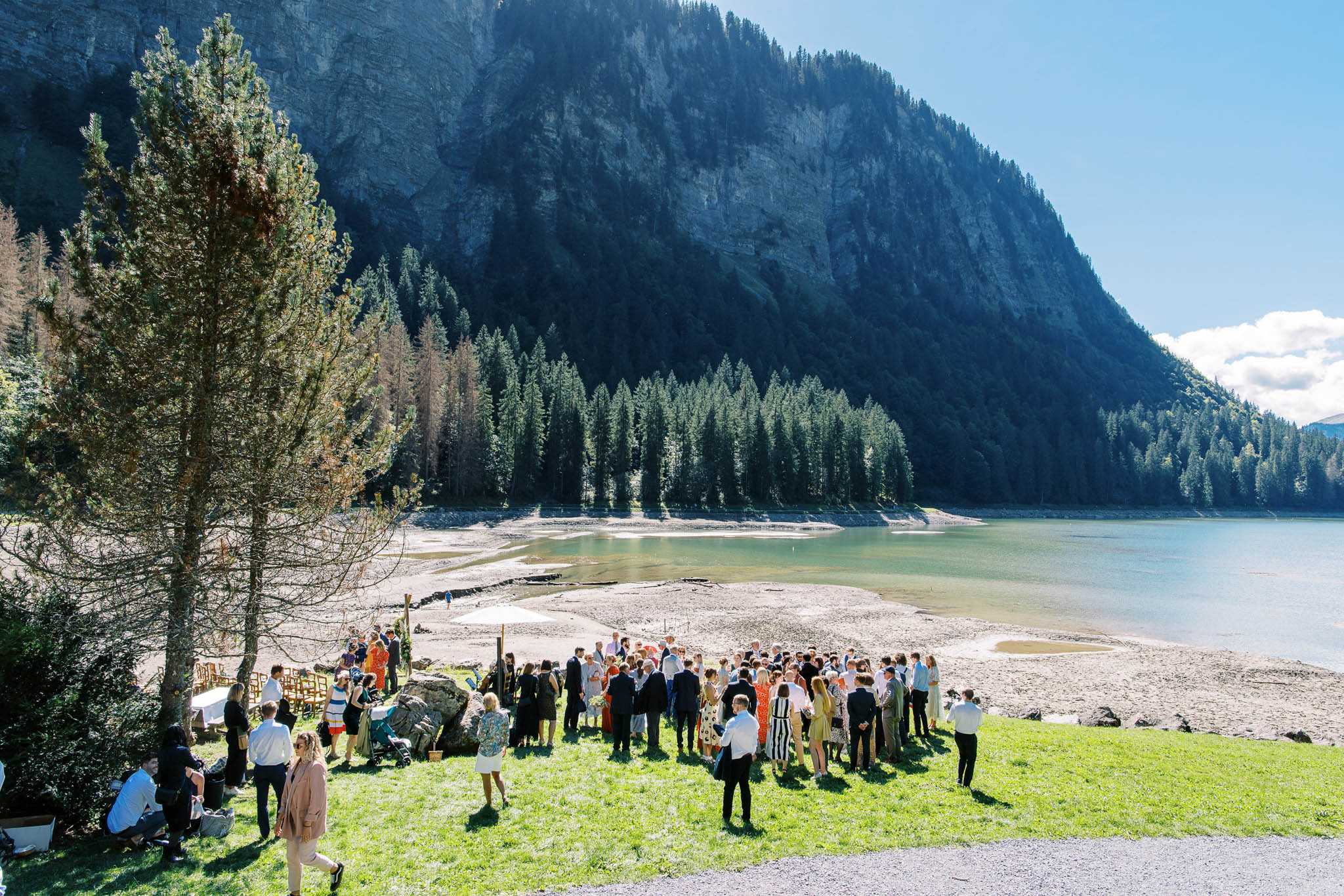 An outdoor wedding ceremony or cocktail hour taking place on the shore of an alpine lake, with approximately 80–100 guests gathered on a grassy area transitioning to a gravel and sandy lakeside. Guests are dressed in a mix of formal and smart-casual attire, including dark suits, colorful summer dresses in stripes, florals, and solids, and a few women in white. A small seating arrangement with wooden chairs and a white umbrella is visible to the left, suggesting a ceremony setup or canapé station. The wide-angle elevated shot captures the full scale of the gathering against the dramatic backdrop of a steep forested mountain face and turquoise alpine lake water. The overall styling is relaxed and natural, with no visible structured floral or decor installations beyond the basic seating.