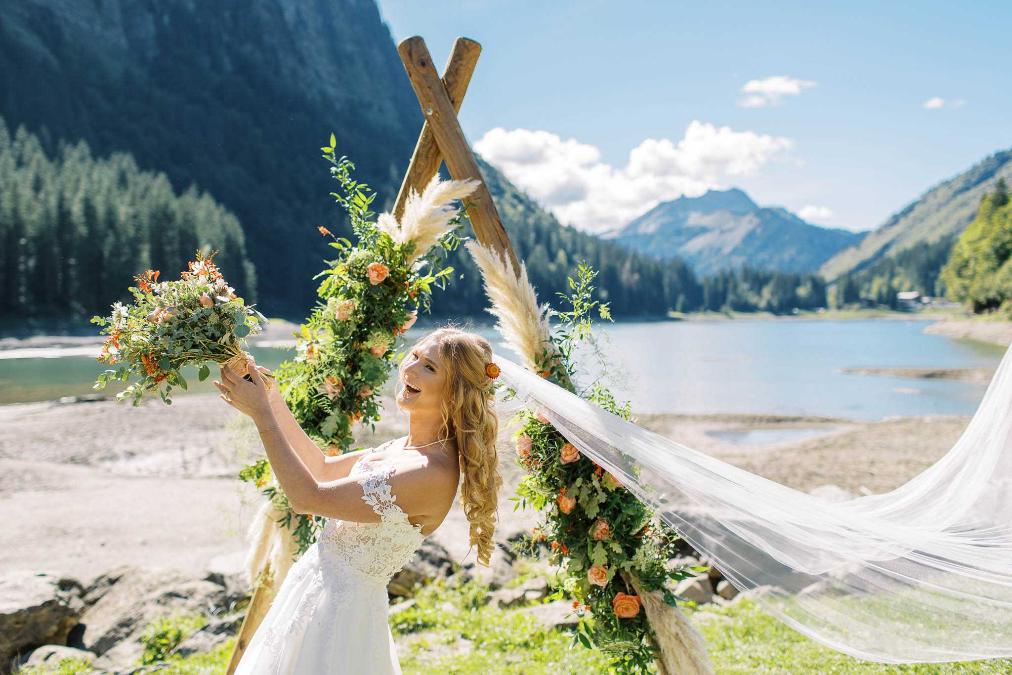 A bridal portrait taken outdoors at a mountain lakeside ceremony setting, with the bride standing in front of a rustic wooden teepee-style arch decorated with lush greenery, peach and orange garden roses, white daisy-like blooms, and pampas grass. The bride wears a white lace bodice gown with a cathedral-length veil billowing dramatically in the wind, and accessorizes with an orange flower hair clip; she holds her bouquet of peach roses, orange blooms, white flowers, and trailing greenery raised in the air while laughing. The decor palette combines peach, burnt orange, and ivory florals with abundant greenery in a boho style. The shot is a mid-length portrait with a wide environmental background showing a mountain lake and forested alpine peaks.