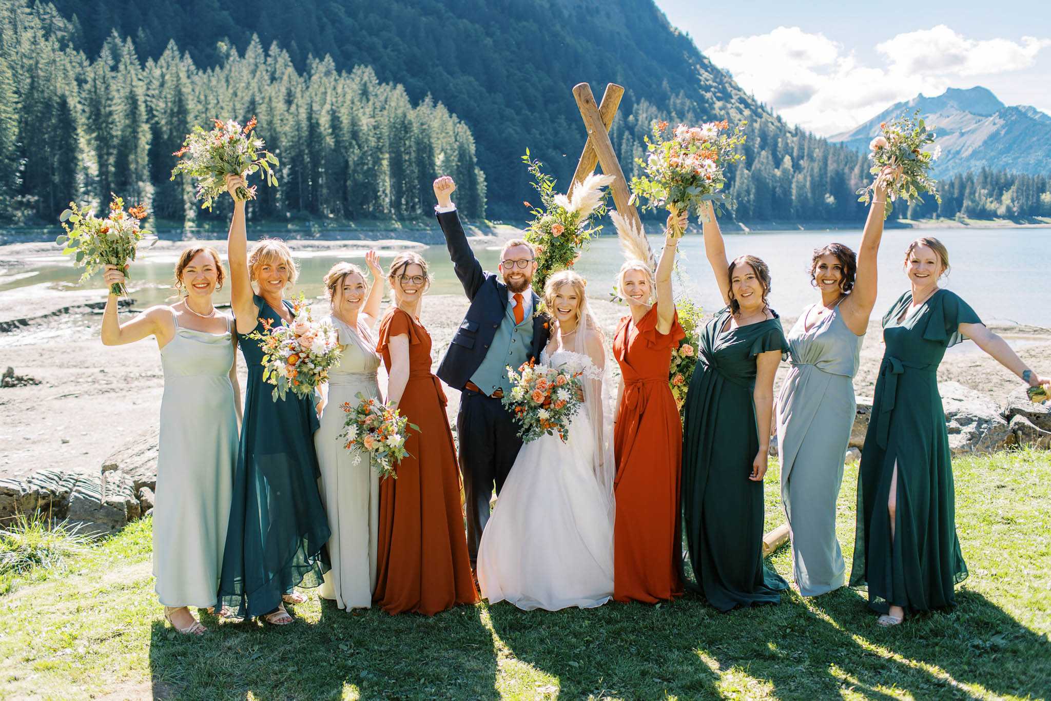 A celebratory bridal party portrait taken outdoors at a lakeside mountain setting, with the couple and eight bridesmaids raising their arms and bouquets in the air. The bride wears a white ballgown with a long veil and holds a bouquet of orange roses, white dahlias, and greenery; the groom wears a navy suit with a light blue vest and an orange tie. The bridesmaids wear mismatched floor-length dresses in a coordinated earthy palette of burnt orange, forest green, dusty sage, and light grey, each holding a loose, organic bouquet of orange and peach blooms with pampas grass and trailing greenery. A rustic wooden cross-style arch decorated with pampas grass and orange florals is visible behind the group. The overall styling theme is boho-rustic with an autumnal color palette.