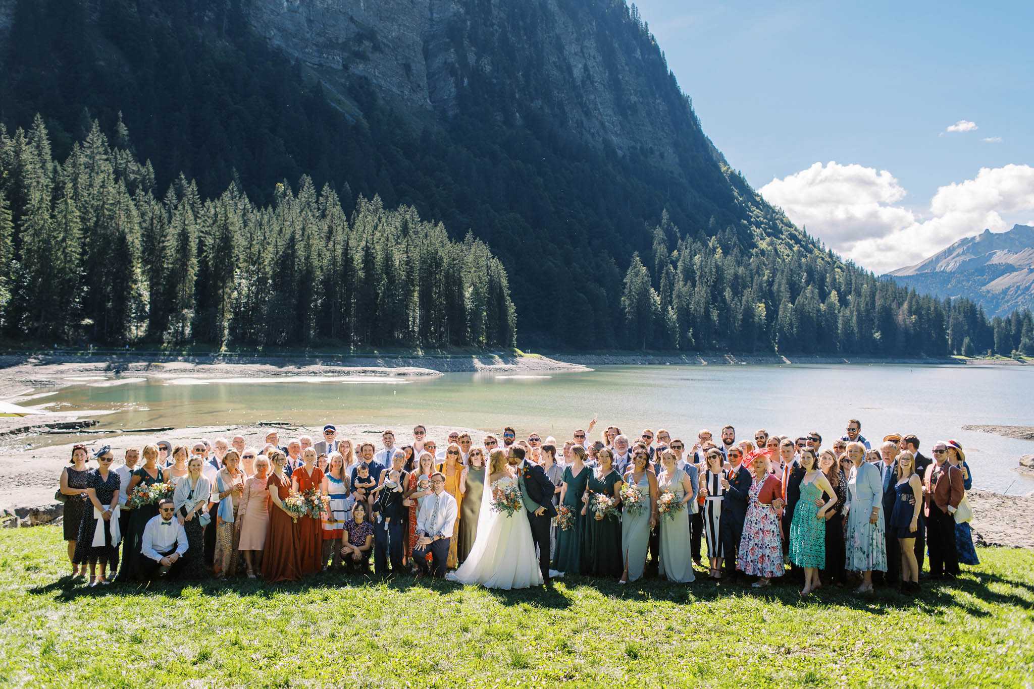 A large group portrait of approximately 80-90 wedding guests gathered outdoors on the shore of an alpine lake, with the bride and groom positioned at the center of the group. The bride wears a white ball gown and the groom is in a dark suit; several bridesmaids in forest green dresses stand nearby holding loose, wildflower-style bouquets in warm tones of rust, peach, and cream. Guests are dressed in a mix of smart-casual and formal attire in varied colors including navy, rust orange, blush, teal, and floral prints, giving the gathering a relaxed, informal feel. The shot is taken from a slightly elevated wide angle, capturing the full group against the backdrop of the lake and steep forested mountain cliffs.
