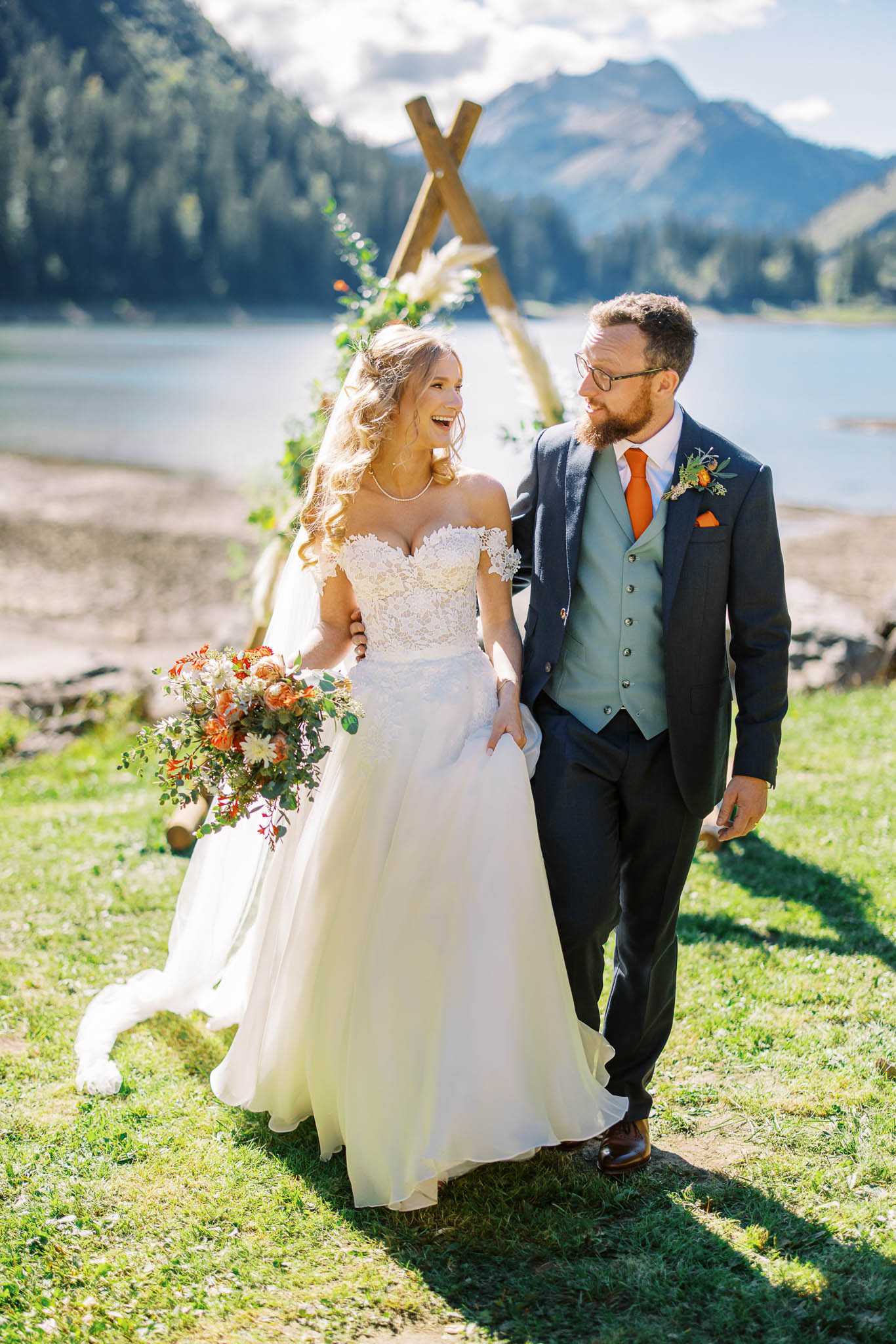 A couple walks together arm-in-arm after their outdoor ceremony beside an alpine lake, with mountains visible in the background. The bride wears an off-the-shoulder ivory lace ballgown with a flowing skirt and carries a large bouquet of burnt orange, rust, and cream blooms with eucalyptus and greenery; the groom wears a charcoal suit with a sage green waistcoat, orange tie, and a matching boutonniere with orange flowers and foliage. Behind them stands a wooden A-frame triangular arch decorated with pampas grass, greenery, and orange florals, reflecting a boho outdoor styling aesthetic. The shot is a medium portrait taken in natural daylight, capturing the bride laughing and the groom looking at her warmly.