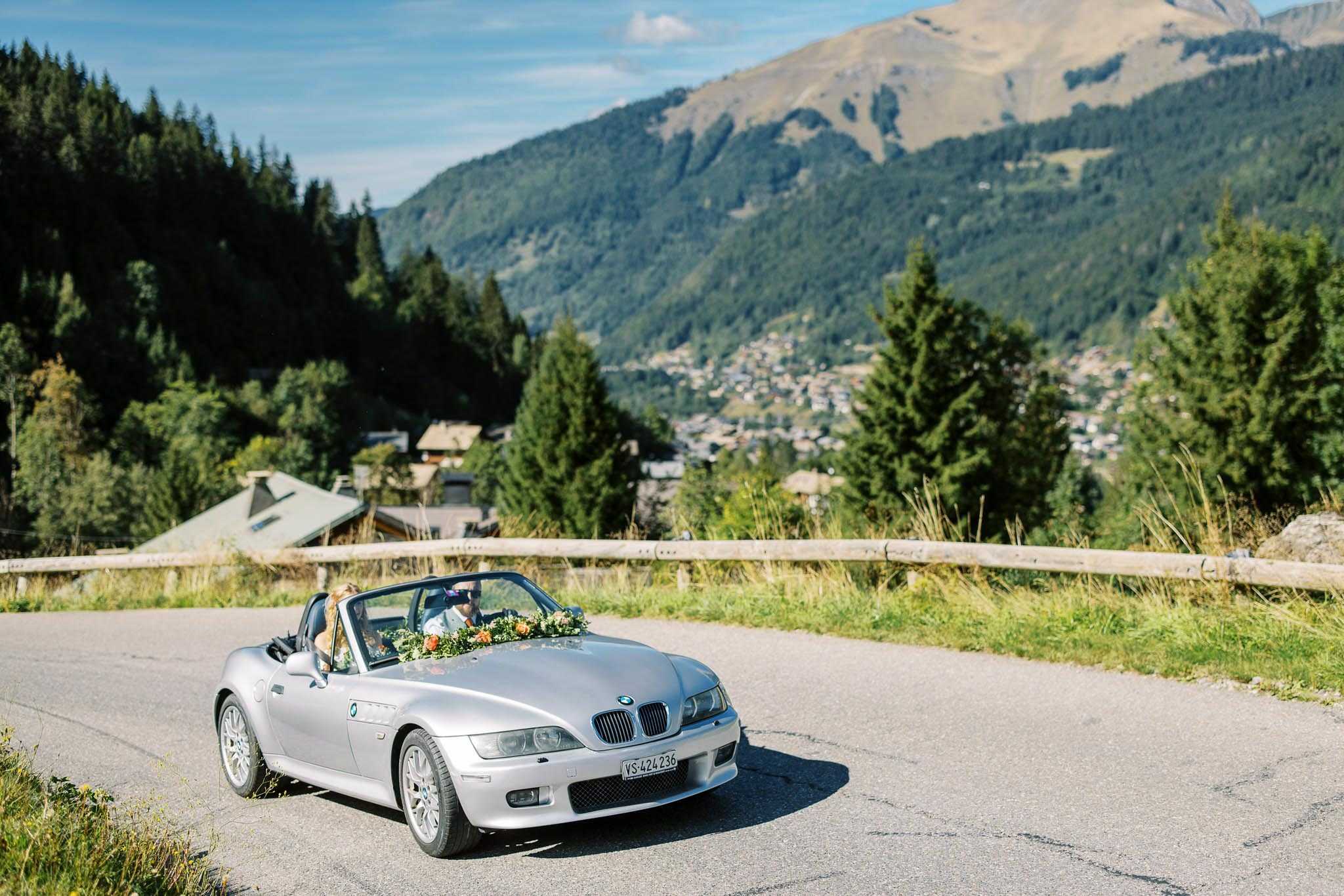 A couple is being transported in a silver BMW Z3 convertible along a mountain road, used as a wedding car. The vehicle is decorated across the windshield with a floral garland featuring orange and peach blooms mixed with greenery. Two occupants are visible — the bride with long blonde hair in the passenger seat and the driver beside her. The wide-angle shot is taken outdoors on a sunny day with an Alpine village and forested mountain peaks visible in the background, suggesting a mountain wedding location in the Alps.