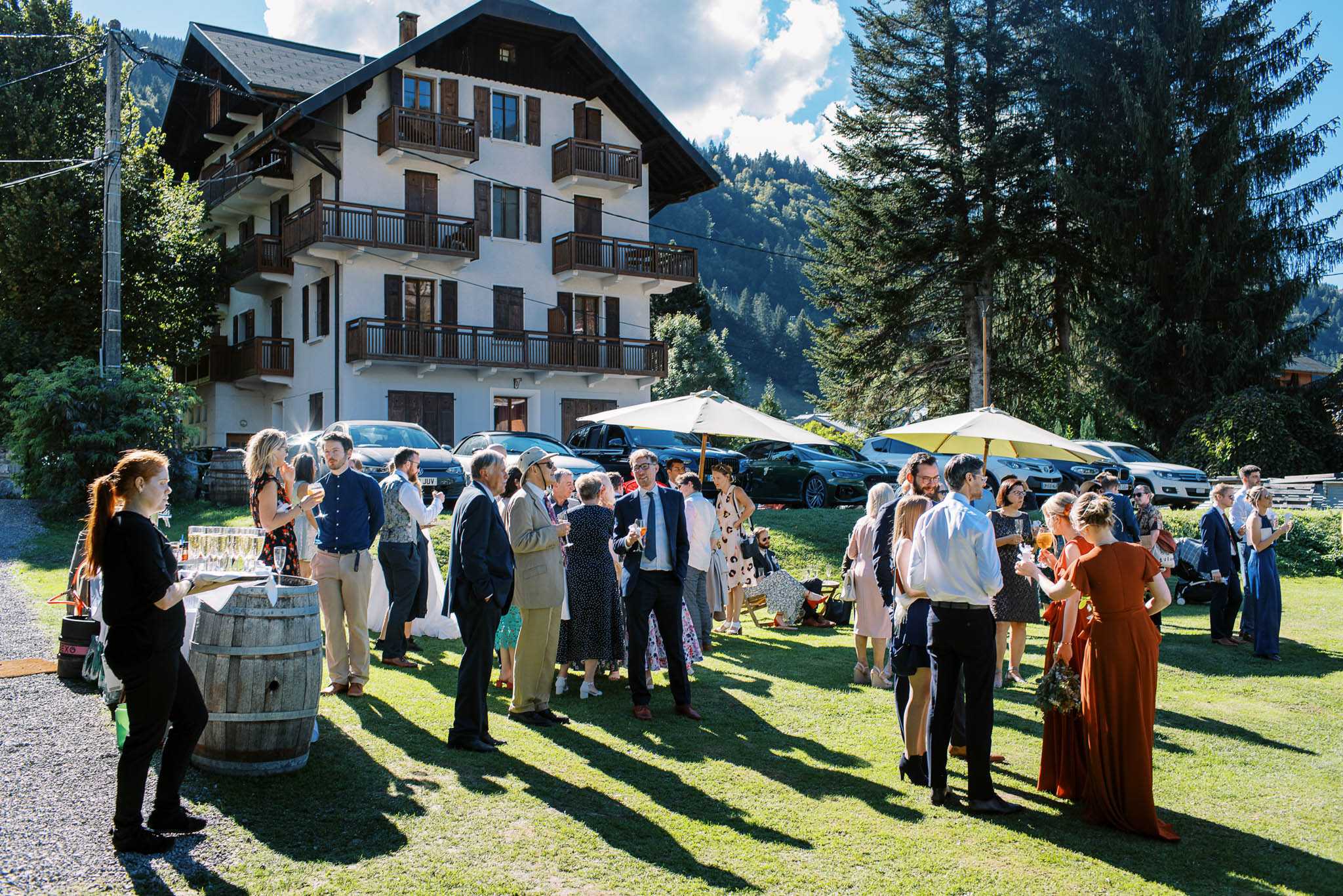 An outdoor cocktail hour taking place on a lawn in front of a large Alpine-style chalet building with white render, dark wood balconies, and shutters, set against a forested mountain backdrop. Approximately 30–40 guests are mingling in small groups, holding glasses of champagne, dressed in smart-casual attire including navy suits, a terracotta floor-length dress, floral dresses, and linen-toned trousers. A wooden barrel serves as a drinks station on the left, topped with a tray of champagne flutes, attended by a staff member in all black; two cream market umbrellas provide shade on the right side of the lawn. The decor styling is relaxed and rustic, incorporating the wine barrel as a functional display piece. Wide shot capturing the full scene including the venue building, guests, and surrounding alpine setting. Potential venue feature image.