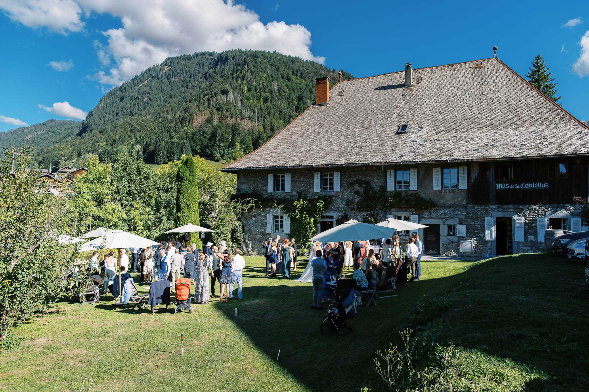 An outdoor cocktail hour taking place on the lawn of Mas de la Coutettaz, a large traditional Alpine stone farmhouse with a steeply pitched slate roof, white shuttered windows, and climbing ivy on the facade. Approximately 40-50 guests are mingling across the grass, dressed in smart-casual summer attire including floral dresses, navy and teal outfits, and light-colored shirts; a bride in a white dress is visible among the crowd. Several large cream market umbrellas are set up across the lawn providing shade, alongside small dark-linen-covered bar or drinks tables. The wide-angle shot captures the full venue exterior and surrounding grounds, with a forested mountain rising behind the property. Potential venue feature image.