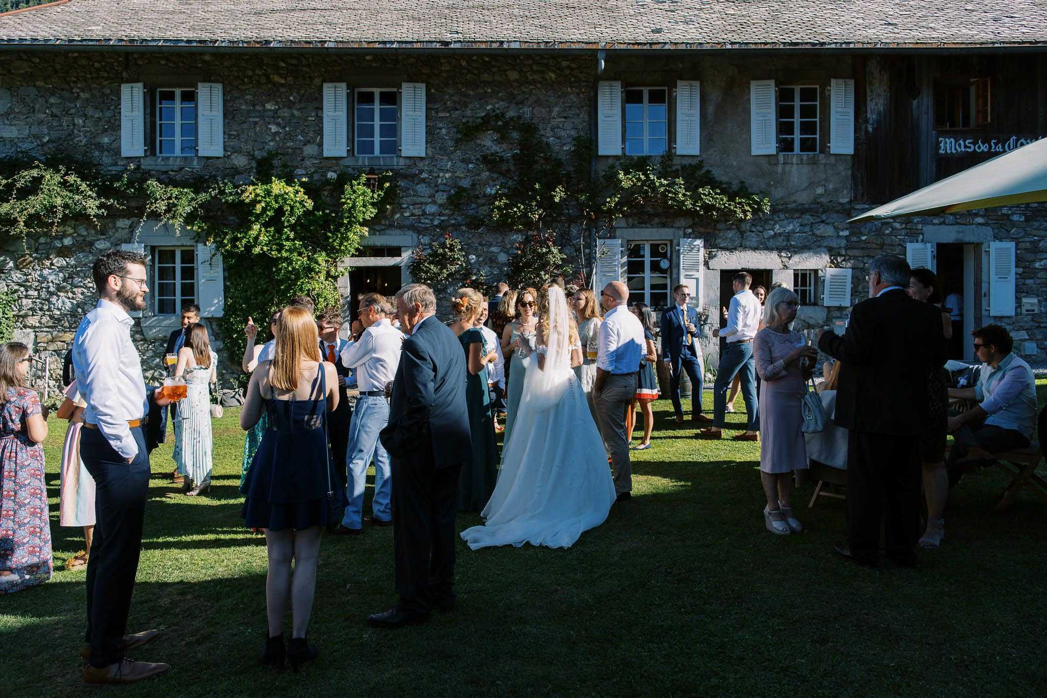 An outdoor cocktail hour taking place on the lawn of a large two-story stone farmhouse with white shutters, partially covered in climbing vines, with a sign partially visible reading 'Mas de la...' The bride, wearing a white gown with a cathedral-length veil and a train, stands with her back to the camera mingling with approximately 25–30 guests. Guests are dressed in smart-casual to semi-formal attire including navy, teal, and floral dresses, with several men in dress shirts and suits; some are holding drinks including what appears to be beer and champagne. The wide-angle shot captures the full facade of the stone venue building alongside the gathered crowd in bright daytime sunlight. Potential venue feature image.