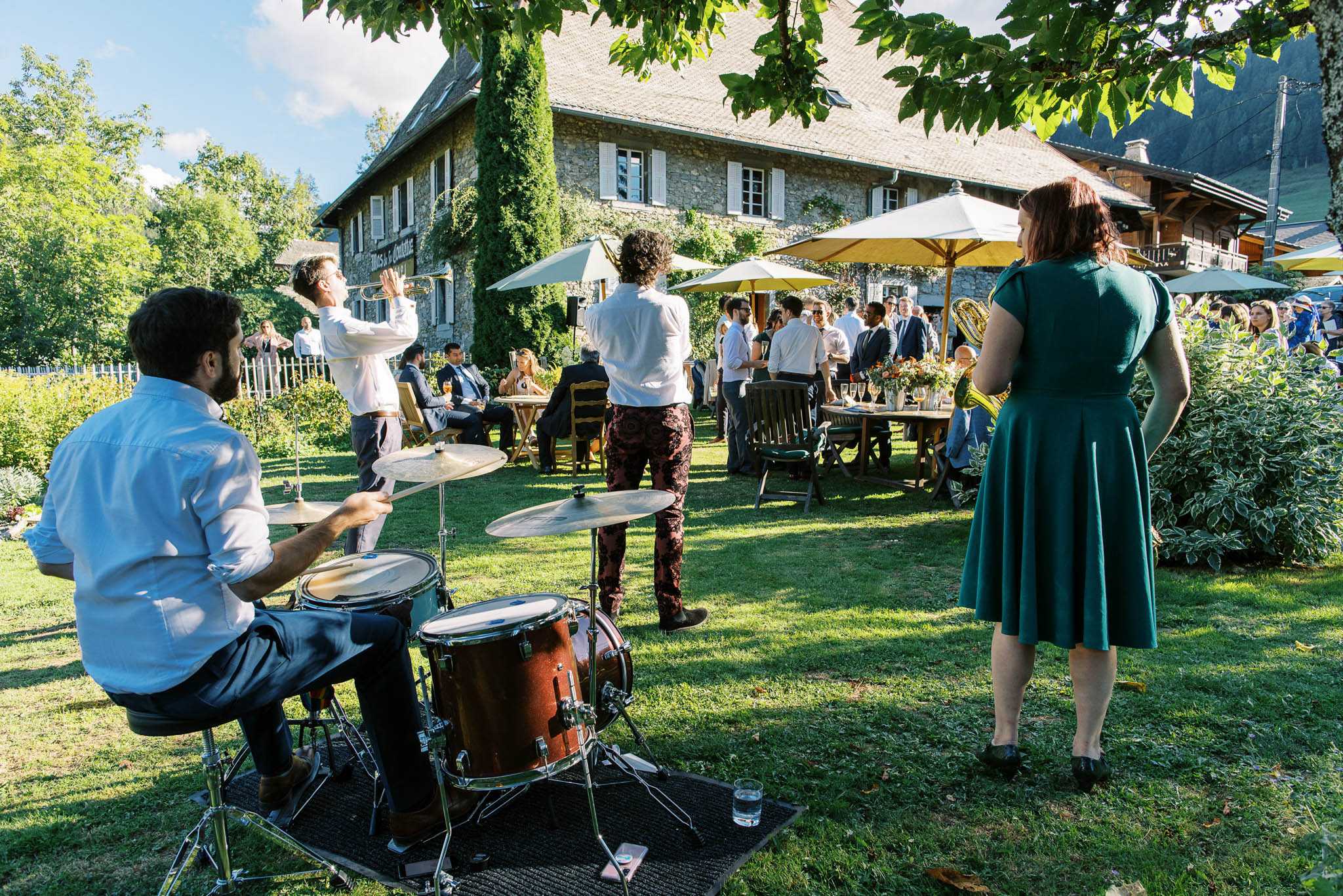 An outdoor cocktail hour taking place on the lawn of an ivy-covered stone farmhouse venue, with approximately 30–40 guests mingling around wooden tables and chairs shaded by yellow and white market umbrellas. In the foreground, a live band performs on the grass, with a drummer seated at a brown drum kit, a trumpet player in a white shirt, and a second musician in dark floral-patterned trousers; a woman in a teal green mid-length swing dress stands nearby watching the performance. Floral centerpieces with orange and warm-toned blooms decorate the tables, and guests are dressed in smart-casual and formal attire. The wide-angle shot captures both the band setup in the foreground and the full party scene extending toward the rustic stone building in the background. Potential venue feature image.