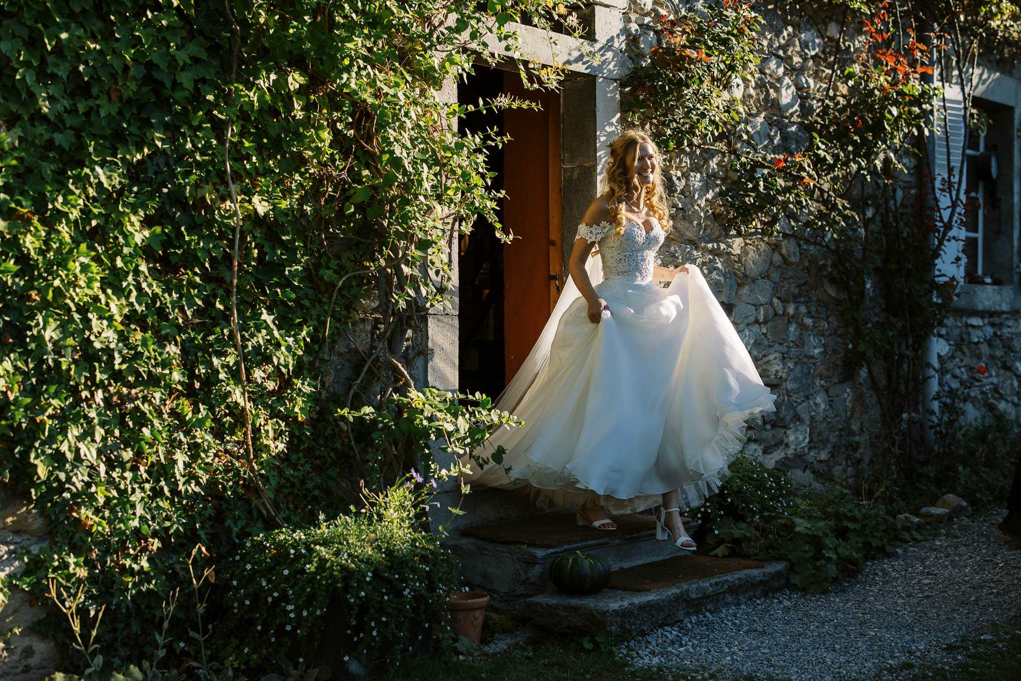 A bride steps out of a doorway of a stone building covered in climbing vines, lifting the skirt of her ivory off-the-shoulder ballgown with a lace-embroidered bodice as she descends stone steps. She is laughing, wearing white block-heeled sandals and has long wavy blonde hair with a floral hair accessory. The setting is outdoor, in front of a rustic stone-walled building with a warm-toned wooden door, surrounded by dense greenery. This is a full-length environmental portrait shot in natural dappled sunlight with a classic-meets-rustic styling feel.