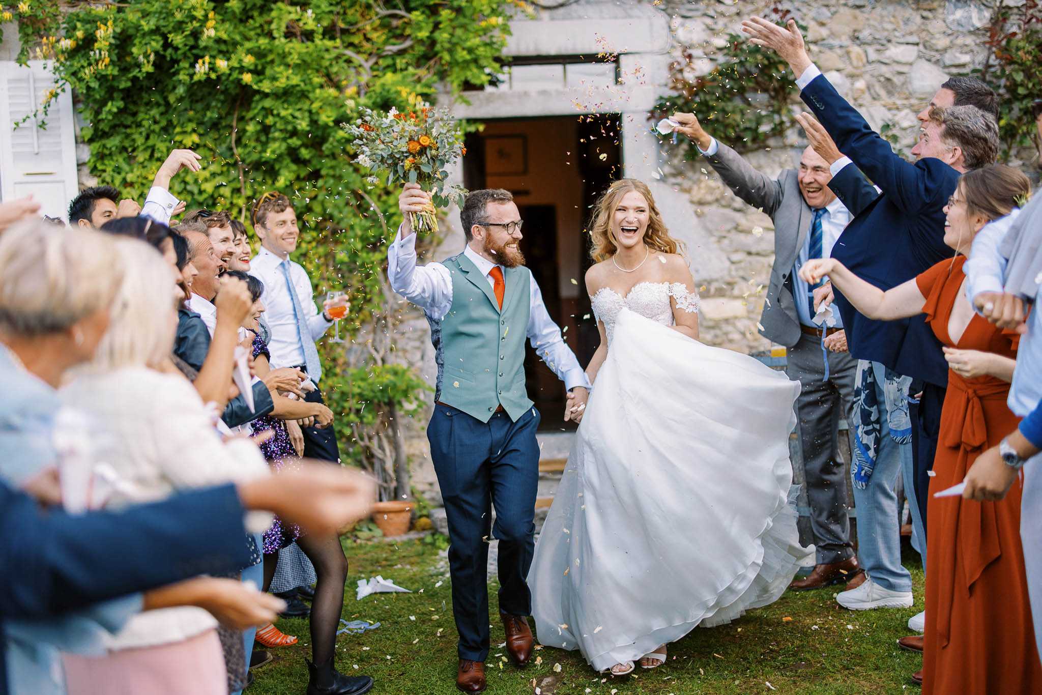 The bride and groom walk through a confetti exit tunnel formed by approximately 20 guests outside a stone building with vine-covered walls. The bride wears an off-the-shoulder white lace ballgown and a pearl necklace, holding her skirt with one hand while the groom raises her bouquet of burnt orange flowers and eucalyptus with the other. The groom wears navy trousers, a sage green waistcoat, a white shirt, and an orange tie with brown leather shoes. Guests are dressed in a mix of navy suits, grey suits, and casual smart attire including a rust-orange wrap dress. The setting is outdoor on a grass lawn in front of a rustic stone building, suggesting a French countryside property. The shot is a wide, eye-level action photograph capturing the energy and movement of the moment, with confetti visible mid-air throughout the frame.