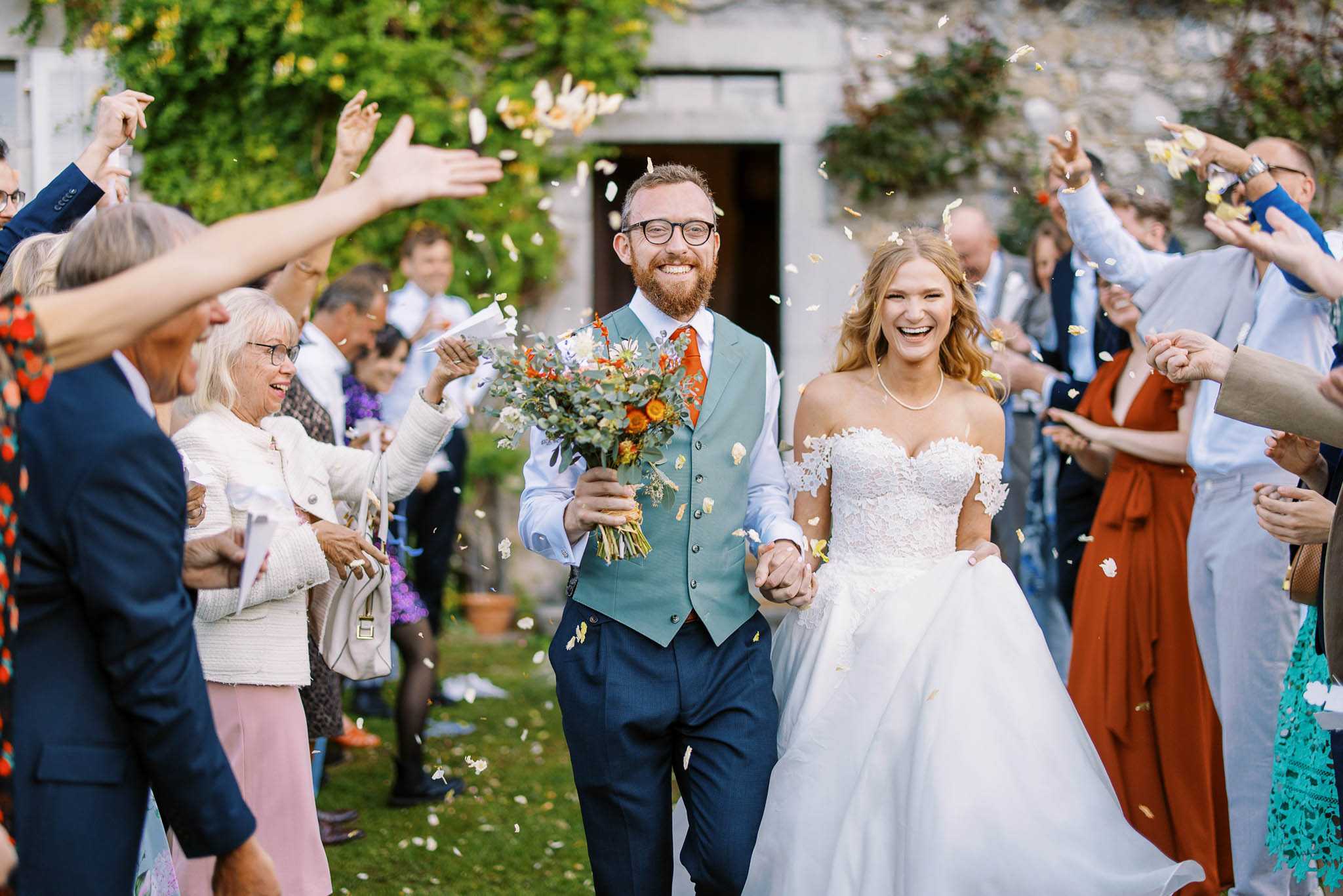 A confetti exit moment captured outdoors in front of a stone building, with the newlywed couple walking hand-in-hand through two lines of approximately 20–25 cheering guests tossing white and ivory petal confetti. The bride wears an off-the-shoulder white lace ballgown with a sweetheart neckline and a pearl necklace, while the groom wears navy trousers, a teal/sage waistcoat, a light blue shirt, and a burnt orange tie; he carries the bridal bouquet, which features orange ranunculus, white daisies, and abundant eucalyptus foliage. Both are laughing and visibly joyful, with guests in a colorful mix of navy suits, a rust-red dress, teal lace, and white tweed also smiling and celebrating. The shot is a medium wide portrait taken at eye level, with the couple sharp in the foreground and guests slightly motion-blurred on either side.