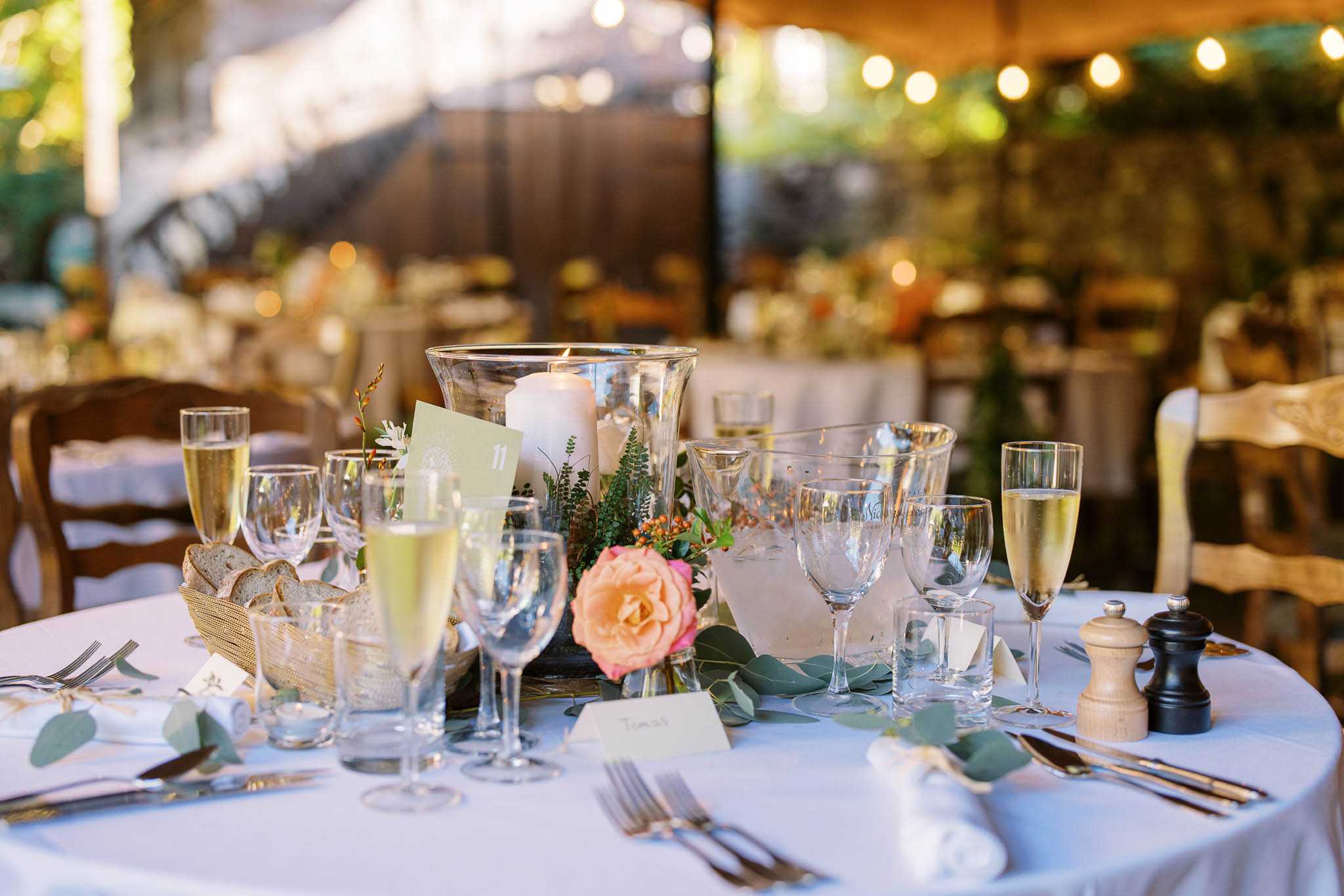 Close-up detail shot of a wedding reception table set in what appears to be a semi-covered outdoor venue with warm globe string lights visible in the background. The round table is dressed with a white linen and set with multiple crystal glasses including champagne flutes filled with a pale yellow drink, wine glasses, and water glasses, along with silver cutlery and white folded napkins. The centerpiece features a large glass hurricane vase holding a white pillar candle, surrounded by a low floral arrangement of coral-peach garden roses, small orange berries, fern fronds, and eucalyptus sprigs. A sage green table number card (numbered 11) is tucked into the centerpiece, and individual white place cards are visible at each setting. A wicker bread basket with sliced bread and wooden salt and pepper mills complete the table styling. The overall decor palette is white, coral, and green with a relaxed garden-party aesthetic. Additional set tables are visible but blurred in the background.