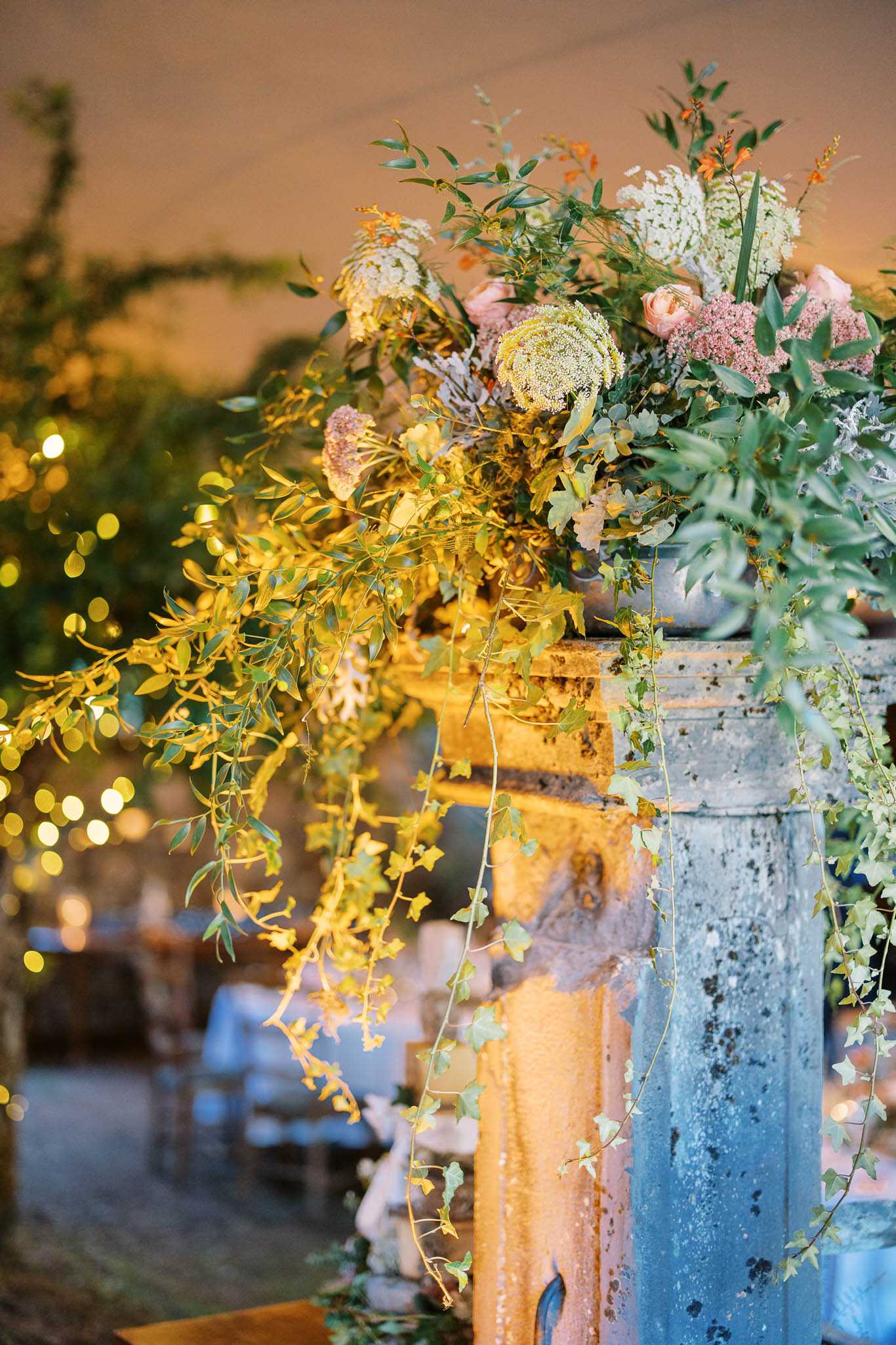 Close-up detail shot of a floral arrangement placed atop a weathered stone balustrade or pillar, likely marking the entrance to an outdoor or semi-covered reception area. The arrangement features a wild, garden-style composition with queen anne's lace, blush garden roses, dusty pink clusters, white blooms, ornamental kale, trailing ivy, eucalyptus, and cascading yellow-green foliage. Warm amber uplighting illuminates the aged stone pillar, while soft bokeh in the background reveals a reception setup with white-linened tables, chairs, and strings of fairy lights. The overall decor palette is organic and garden-inspired, mixing warm and cool tones in a lush, unstructured style.