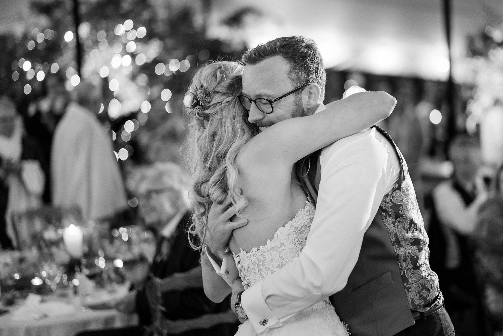 This black-and-white image captures the couple's first dance at an indoor evening reception. The bride, wearing a lace-back dress with loose wavy hair adorned with small floral hair accessories, embraces the groom closely as he holds her around the waist. The groom wears a waistcoat over a white dress shirt with a patterned paisley jacket sleeve visible, and round-framed glasses. The background is softly blurred, showing circular bokeh from string lights or fairy lights overhead, seated guests at candlelit tables with glassware visible to the left, and what appears to be a marquee or tent reception space. The image is a medium close-up portrait shot with strong contrast between light and shadow tones, emphasizing the couple in the foreground against the diffused reception backdrop.