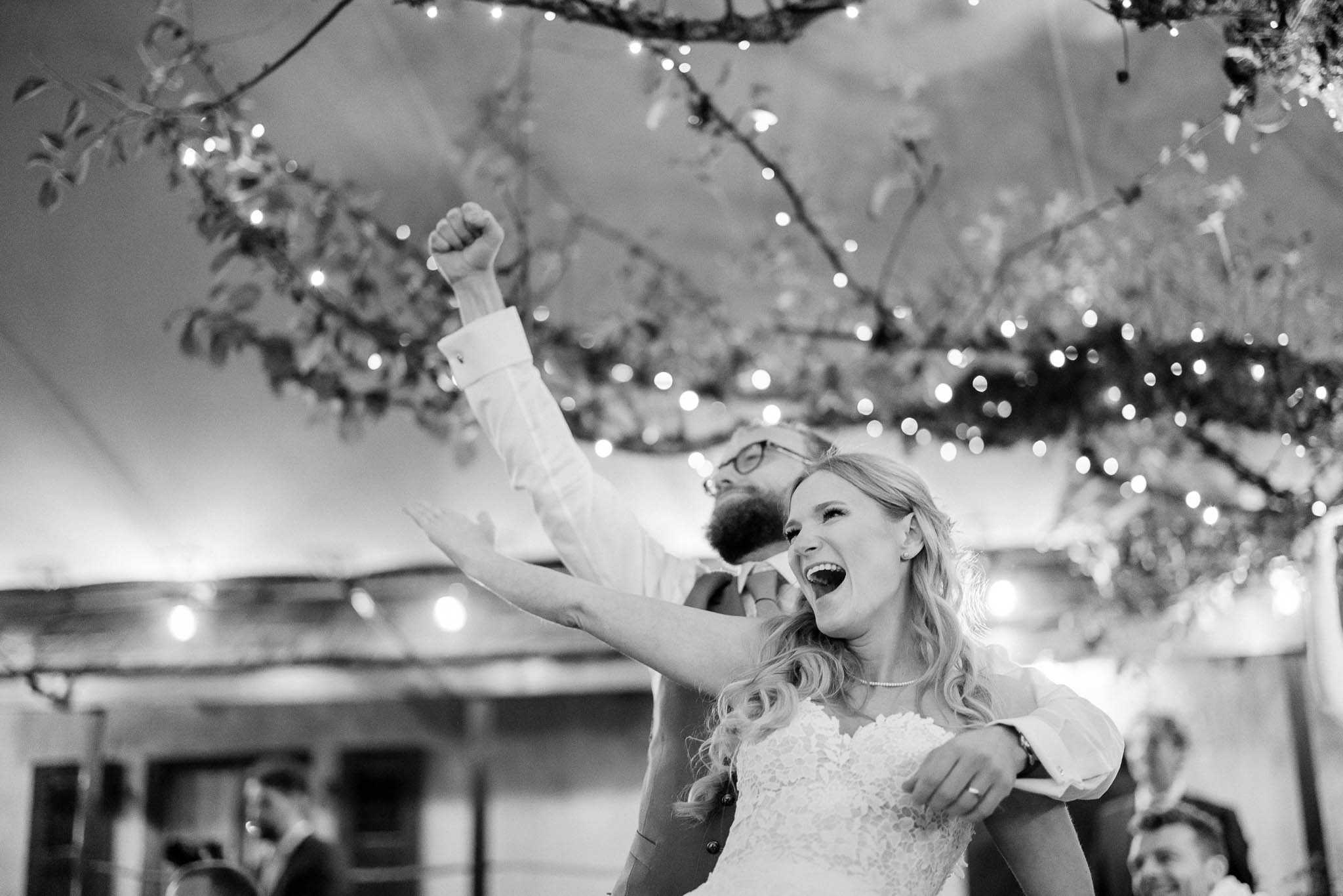 This black-and-white image captures the bride and groom celebrating energetically on the dance floor during the wedding reception. The bride, wearing a strapless lace gown and a delicate pearl necklace, is laughing with her mouth open wide while the groom, in a dress shirt and glasses with a beard, pumps his fist in the air behind her. The reception appears to be held under a large tent or marquee, decorated with fairy lights and branches with foliage forming an overhead canopy installation, creating soft bokeh light throughout the frame. A few guests are visible in the blurred background, and the composition is a mid-shot taken from a low angle, emphasizing the couple's joyful energy against the lit canopy above.