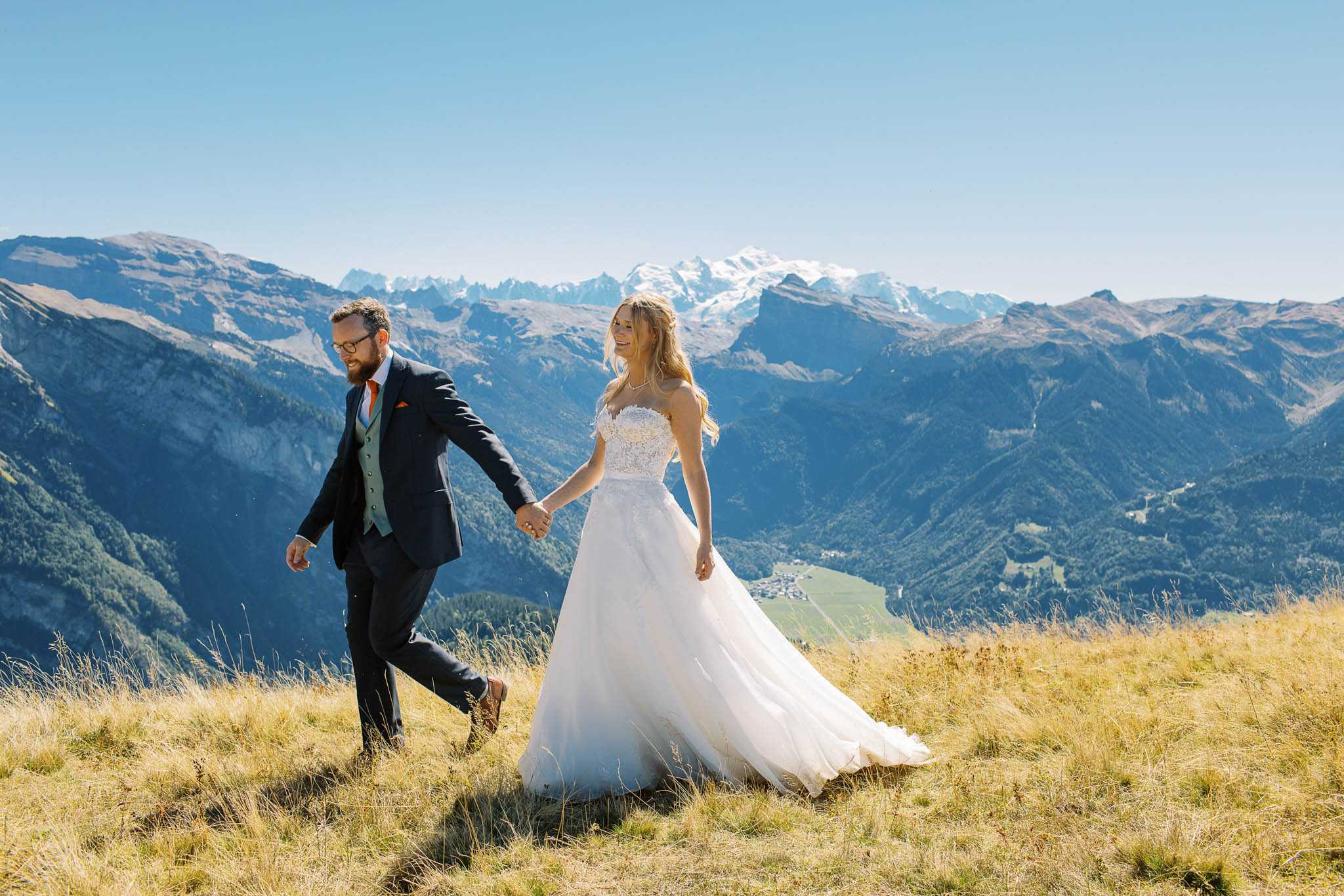 A couple portrait taken outdoors on a high-altitude alpine meadow, with a sweeping mountain range and snow-capped peaks visible in the background. The bride wears a white strapless ballgown with a lace-embroidered bodice and a full tulle skirt, with loose blonde hair and a delicate necklace; the groom wears a dark navy suit with a sage green waistcoat, an orange tie, and a matching pocket square. The two are walking hand-in-hand across dry golden-toned grass, with the bride smiling and looking back over her shoulder. The shot is a mid-length portrait taken in bright natural daylight with a wide-angle composition that emphasizes the dramatic alpine backdrop.