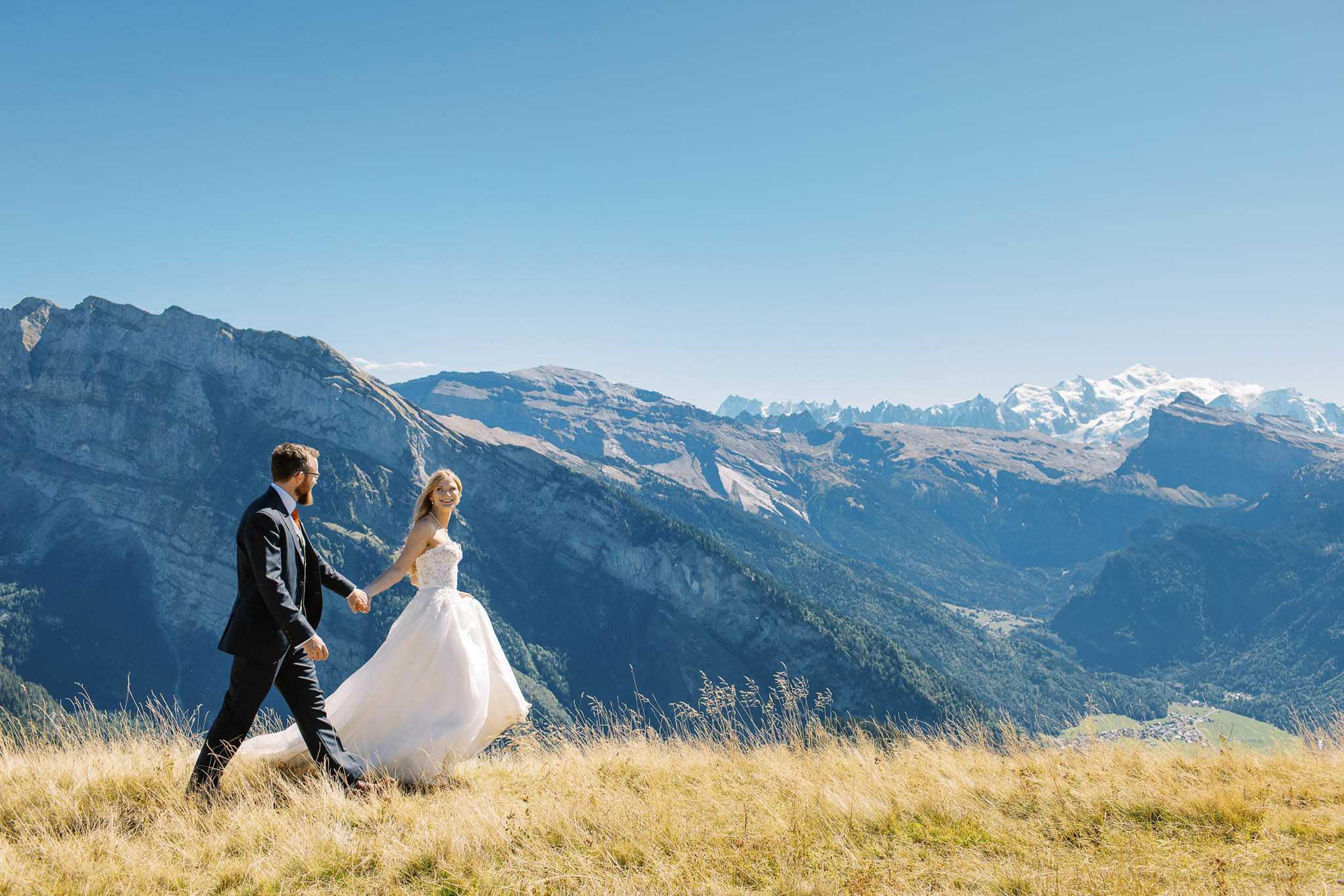 A couple portrait session set outdoors on a high-altitude mountain ridge, with an expansive alpine valley and snow-capped peaks visible in the background. The bride wears a strapless ivory ballgown with a lace-appliqué bodice and a full tulle skirt that billows in the wind, while the groom wears a dark navy suit with an orange tie. The two are walking hand-in-hand across dry golden grass, with the bride looking back toward the camera smiling and the groom looking ahead. Wide shot composition capturing both the couple and the dramatic mountain landscape in full.