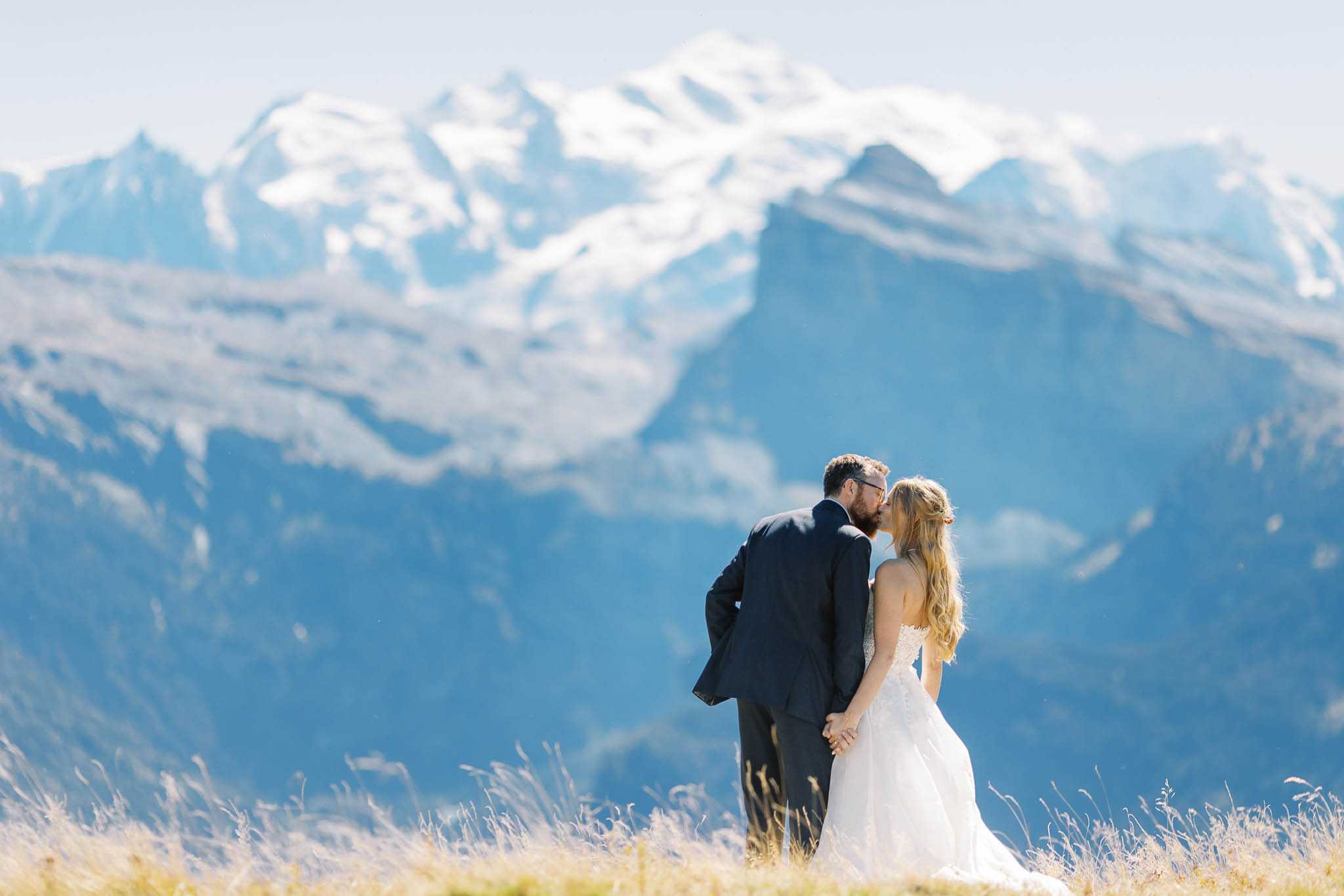 A couple shares a kiss during an outdoor wedding portrait session on an alpine hillside, with snow-capped mountain peaks visible in the background. The bride wears a strapless white ballgown with lace detailing and has long wavy blonde hair, while the groom wears a dark charcoal suit. The two are holding hands, positioned centrally in the frame with the vast mountain range softly blurred behind them. This is a wide-shot couple portrait taken in natural daylight, with the shallow depth of field drawing focus to the pair against the dramatic alpine backdrop.