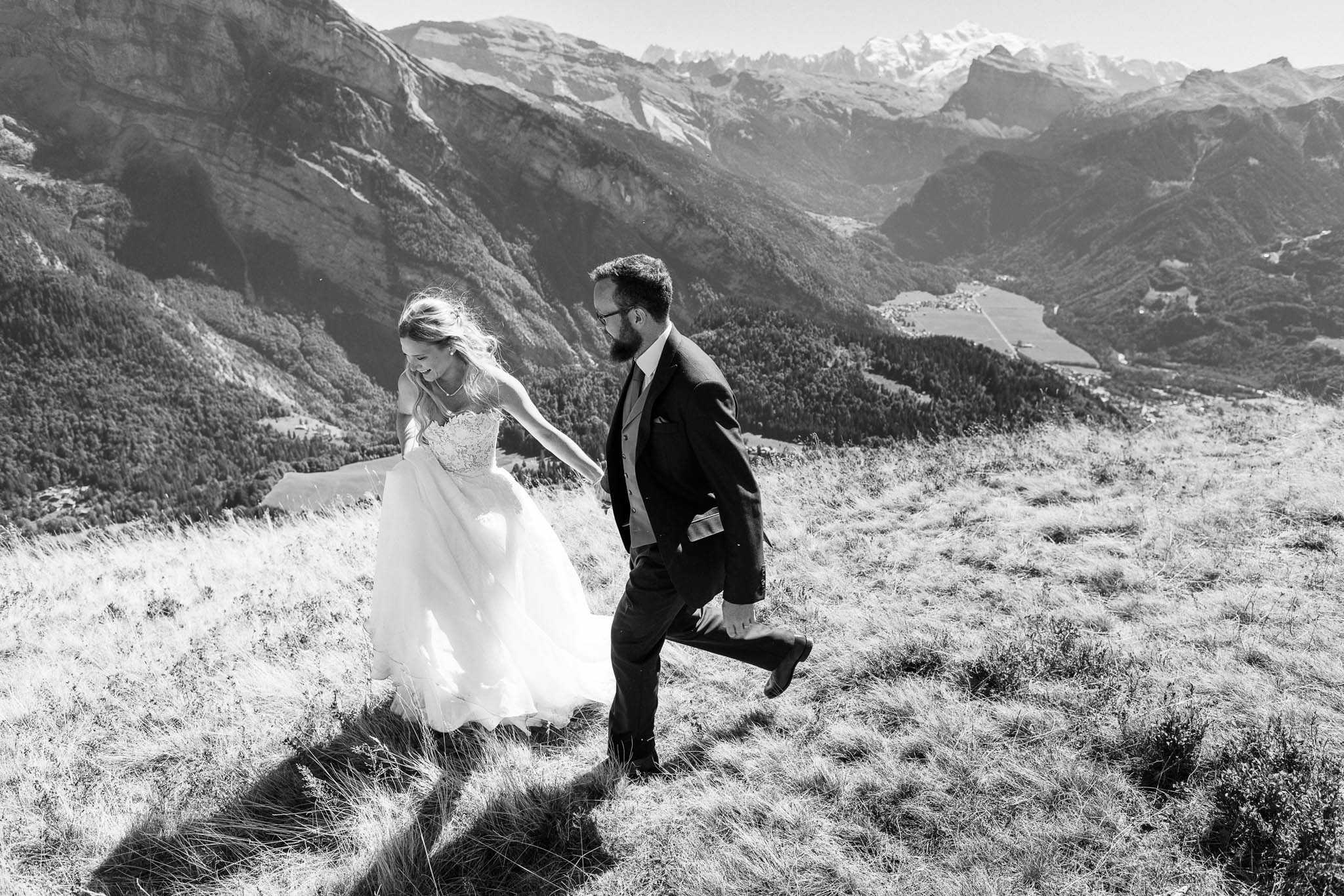 Black-and-white couple portrait taken outdoors on a high-altitude alpine hillside, with a sweeping mountain valley and snow-capped peaks visible in the background. The bride and groom are walking and laughing together up a grassy slope, holding hands — she leads slightly ahead while he looks toward her with a smile. The bride wears a strapless ballgown with a lace bodice and full tulle skirt, her hair loose and windswept; the groom is dressed in a dark suit with a waistcoat, tie, and glasses. The image is high-contrast B&W with bright highlights on the dress and deep shadows across the hillside terrain. Wide environmental portrait shot capturing both the couple and the dramatic mountain landscape behind them.