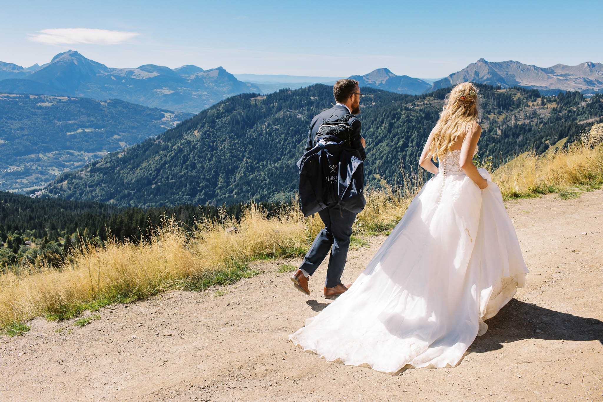 A bride and groom are walking together along a dirt mountain path, photographed from behind in a wide shot. The setting is an outdoor alpine location at high elevation, with a panoramic view of forested mountain ranges visible in the background. The bride wears a full-skirted white ballgown with a lace-detailed corset back and a long train trailing on the path, with small red floral accents in her loose wavy blonde hair. The groom wears a navy suit with brown leather shoes and carries a hiking backpack with a jacket attached to it, suggesting an adventurous, destination-style portrait session. The overall styling reflects a relaxed, adventure-elopement aesthetic combined with formal bridal attire.
