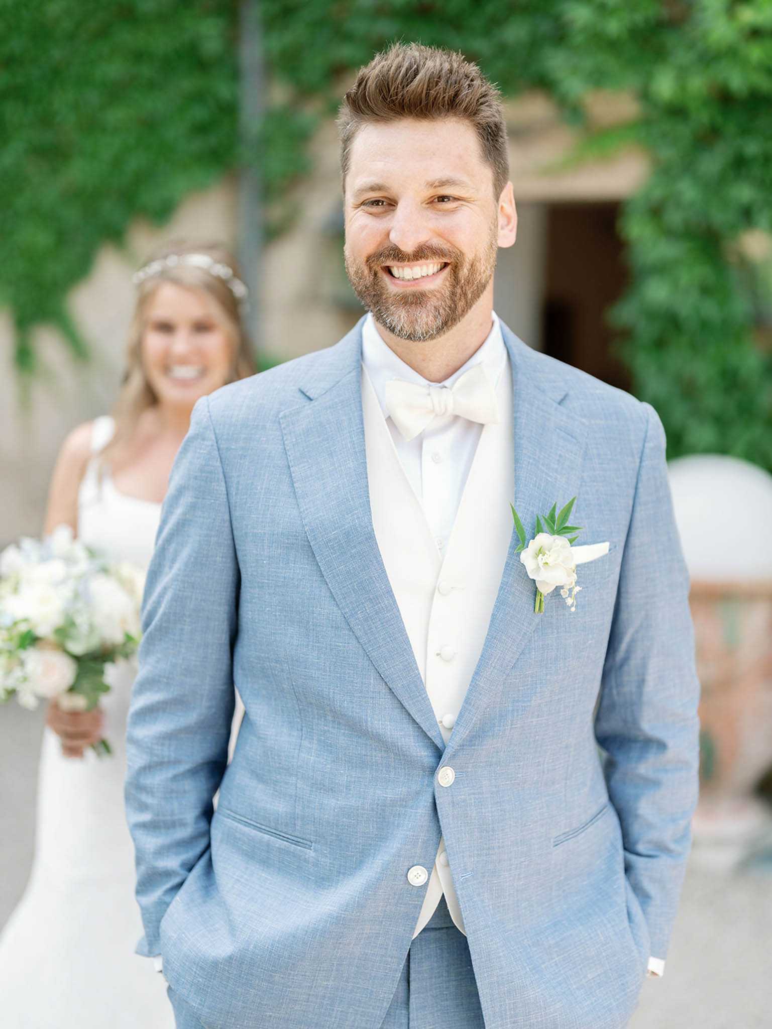 Portrait of a groom standing outdoors in a courtyard setting with ivy-covered stone walls in the background. He is wearing a light blue suit with a cream waistcoat, cream bow tie, and a white floral boutonniere featuring a peony and greenery with small white accent blooms. In the soft-focus background, the bride is visible wearing a white dress and a floral hair crown, holding a white and green bouquet. The composition is a mid-shot portrait focused on the groom, with the bride positioned behind him, suggesting a first-look or post-ceremony moment. The overall styling palette is light blue and ivory with white florals.