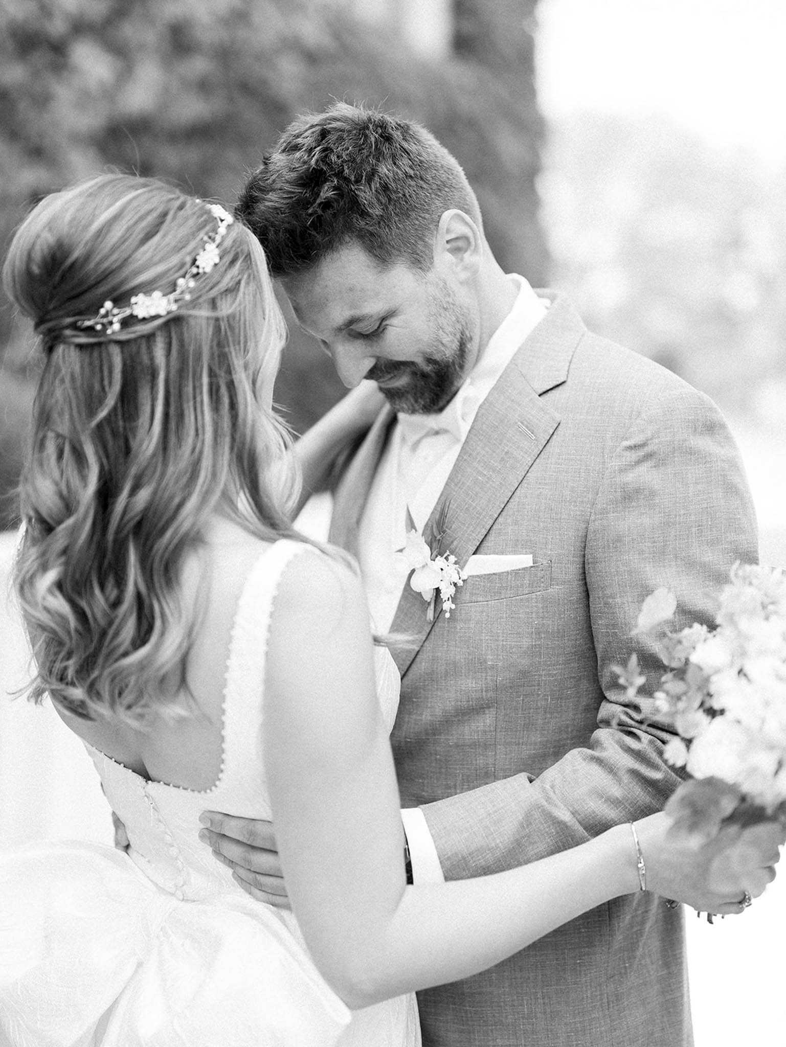This black-and-white couple portrait shows a bride and groom in an intimate embrace outdoors, with soft mid-tones and gentle contrast throughout. The bride, photographed from behind, wears a sleeveless dress with a scalloped back neckline and has her hair styled in loose waves with a delicate floral hair vine accessory; she holds a small bouquet with foliage and small blooms visible at the right edge of the frame. The groom wears a textured light-toned suit with a white pocket square and a small floral boutonniere featuring delicate clustered blooms, and he rests one hand on the bride's waist while looking downward toward her with a smile. The composition is a close-up portrait shot with a blurred natural background.