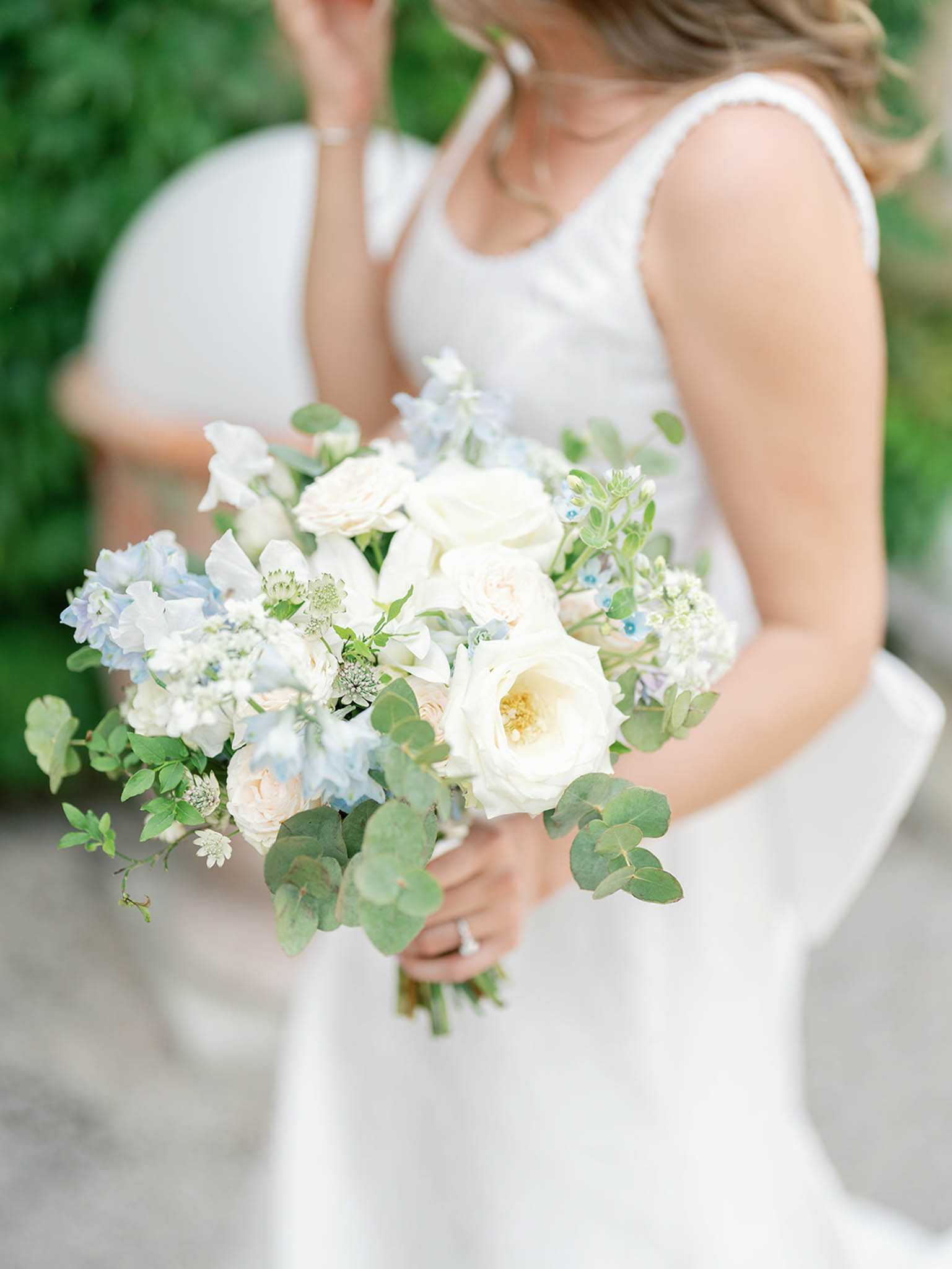 A close-up detail shot of a bride holding her bridal bouquet outdoors, with a second person in white softly blurred in the background. The bride wears a sleeveless white gown with a square or scoop neckline and a diamond ring is visible on her hand. The bouquet is a loosely arranged, garden-style composition featuring cream garden roses, pale blush spray roses, soft blue delphinium, white astrantia, white sweet peas, and trailing eucalyptus stems, creating a soft blue, white, and green color palette.