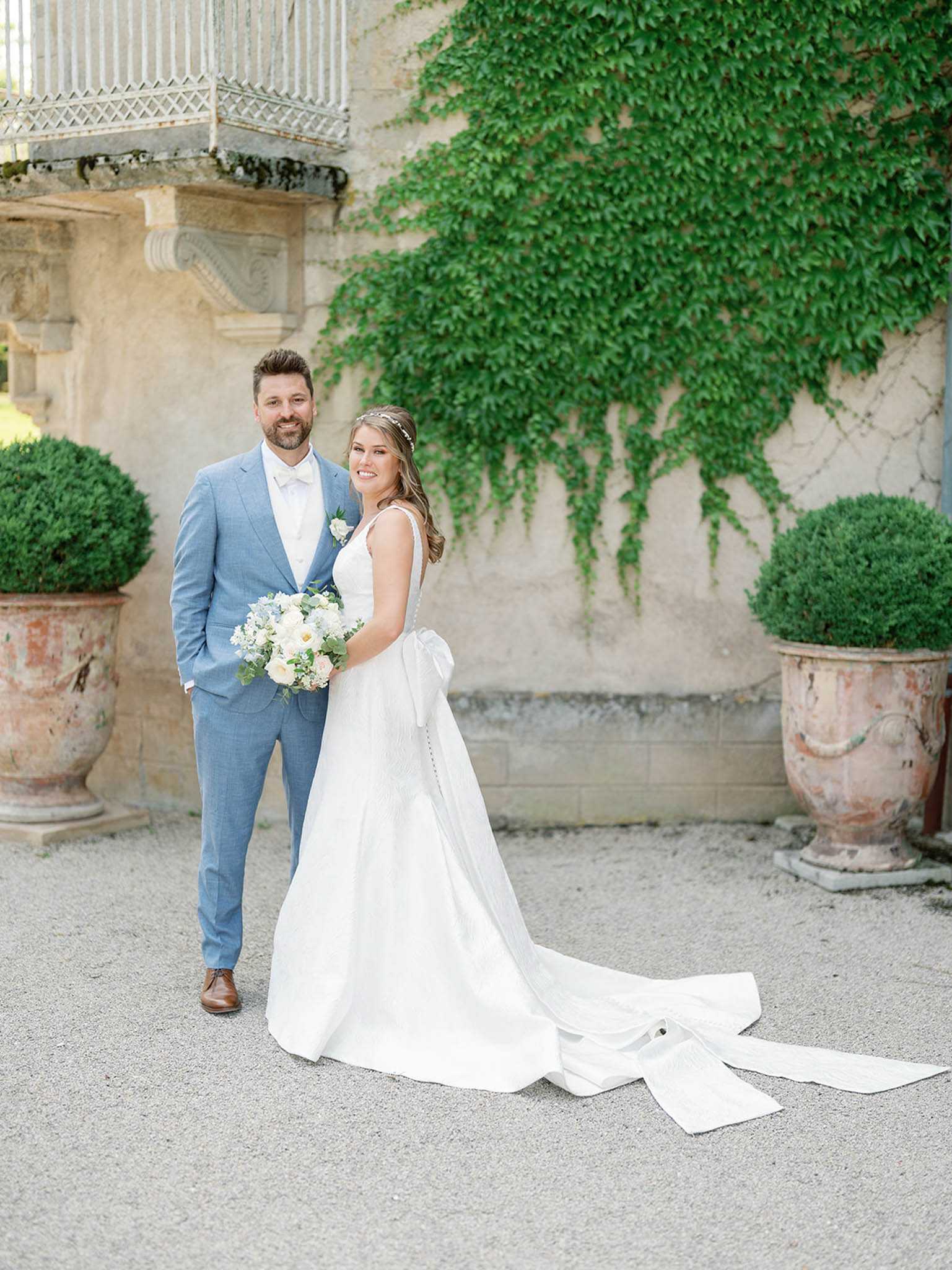 A couple portrait taken outdoors on a gravel courtyard in front of a chateau facade. The bride wears a white A-line gown with a large back bow and an extended train, a delicate floral hairpiece, and carries a bouquet of white peonies, blush roses, dusty blue florals, and eucalyptus. The groom wears a light blue suit with a white tie and a white boutonniere. They stand together facing the camera, flanked by two large aged terracotta urns planted with clipped boxwood topiary spheres. The background features the stone wall of the chateau partially covered in climbing ivy, with a decorative balcony visible in the upper left corner. The shot is a full-length portrait with soft, even natural lighting and a classic French chateau aesthetic.