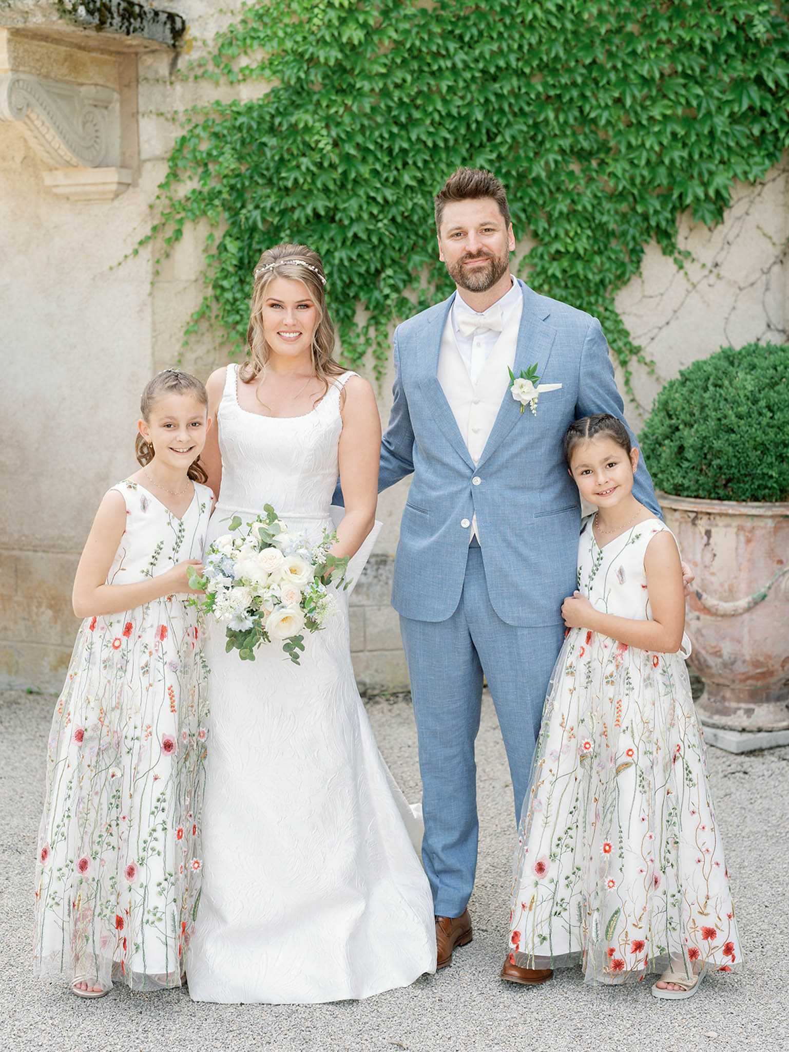A portrait of the bride and groom posing with two flower girls in front of an ivy-covered stone chateau wall, taken outdoors on a gravel courtyard. The bride wears a white A-line jacquard gown with a square neckline and a delicate beaded hair vine, holding a loose bouquet of ivory garden roses, soft blue flowers, and eucalyptus greenery. The groom is dressed in a light blue suit with a white bow tie and a white rose boutonniere with greenery. The two flower girls wear matching white tulle dresses with colorful embroidered wildflower motifs in red, pink, and green. This is a medium full-length group portrait shot.