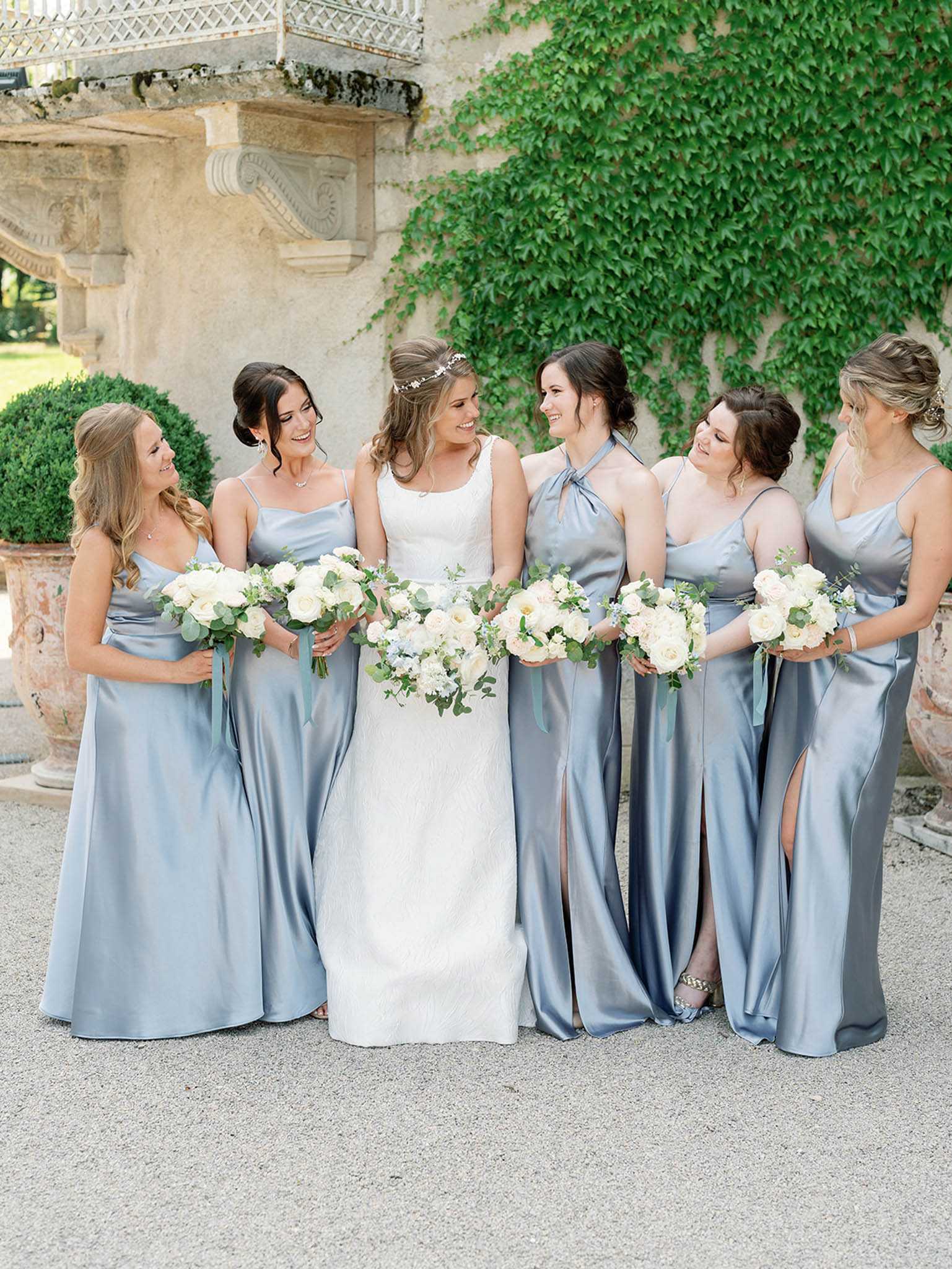 A bridal party portrait taken outdoors against the ivy-covered stone facade of what appears to be a French chateau, with ornate architectural details and a wrought-iron balcony visible in the background. The bride stands at center in a fitted ivory lace sleeveless gown with a square neckline and a delicate floral hair accessory, surrounded by five bridesmaids wearing floor-length steel blue satin dresses in varying styles including spaghetti straps, a halter neck, and a wrap style, several with front slits. Each woman holds a bouquet of ivory and blush garden roses with eucalyptus and dusty blue ribbon streamers, and the group is laughing and looking at one another in a candid moment. The shot is a medium-wide portrait taken at eye level in natural daylight, with a classic French country aesthetic reinforced by the satin color palette and stone chateau backdrop.