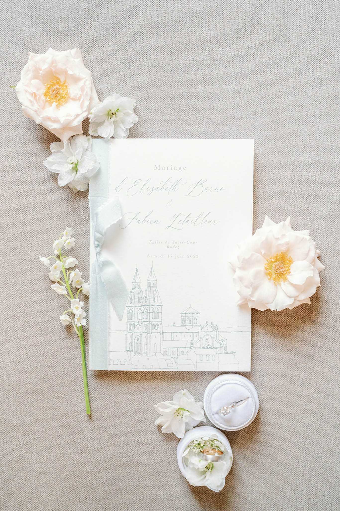 A flat lay detail shot of a French wedding invitation suite for the marriage of Elisabeth Barne and Fabien Letailleur, dated Saturday 17 June 2023 at Église du Sacré-Cœur in Rodez. The ivory ceremony booklet features a sage green line-art illustration of the church facade and is tied with a pale blue-grey silk ribbon bow. Surrounding the stationery are blush garden roses with yellow centers, white delphinium blooms, and a stem of lily of the valley, all arranged on a neutral linen-textured background. Two white velvet ring boxes are positioned at the lower right, one open displaying a diamond solitaire engagement ring and the other containing two gold wedding bands accented with small white florals.