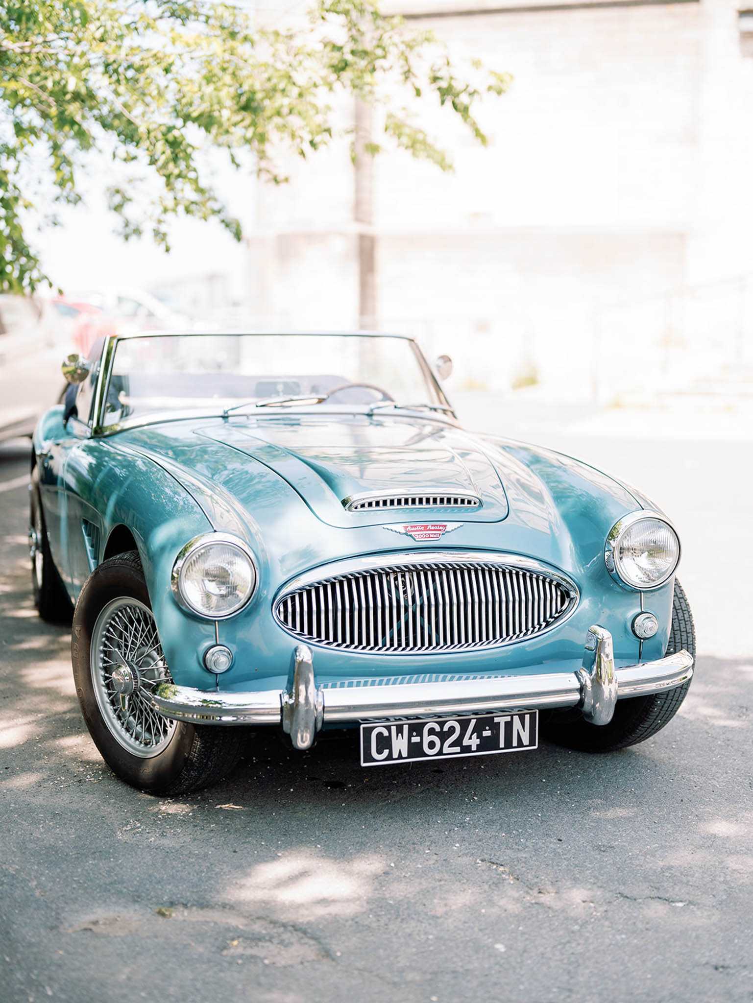 A vintage Austin-Healey 3000 convertible in powder blue with chrome detailing, wire-spoke wheels, and an oval chrome grille is parked on a street, likely used as a wedding transportation vehicle. The car bears a French license plate (CW-624-TN) and has a red interior visible above the windscreen. No people are present in the frame. The shot is a close-up portrait-style composition taken from a slightly low angle at the front of the car, with a softly blurred urban background.